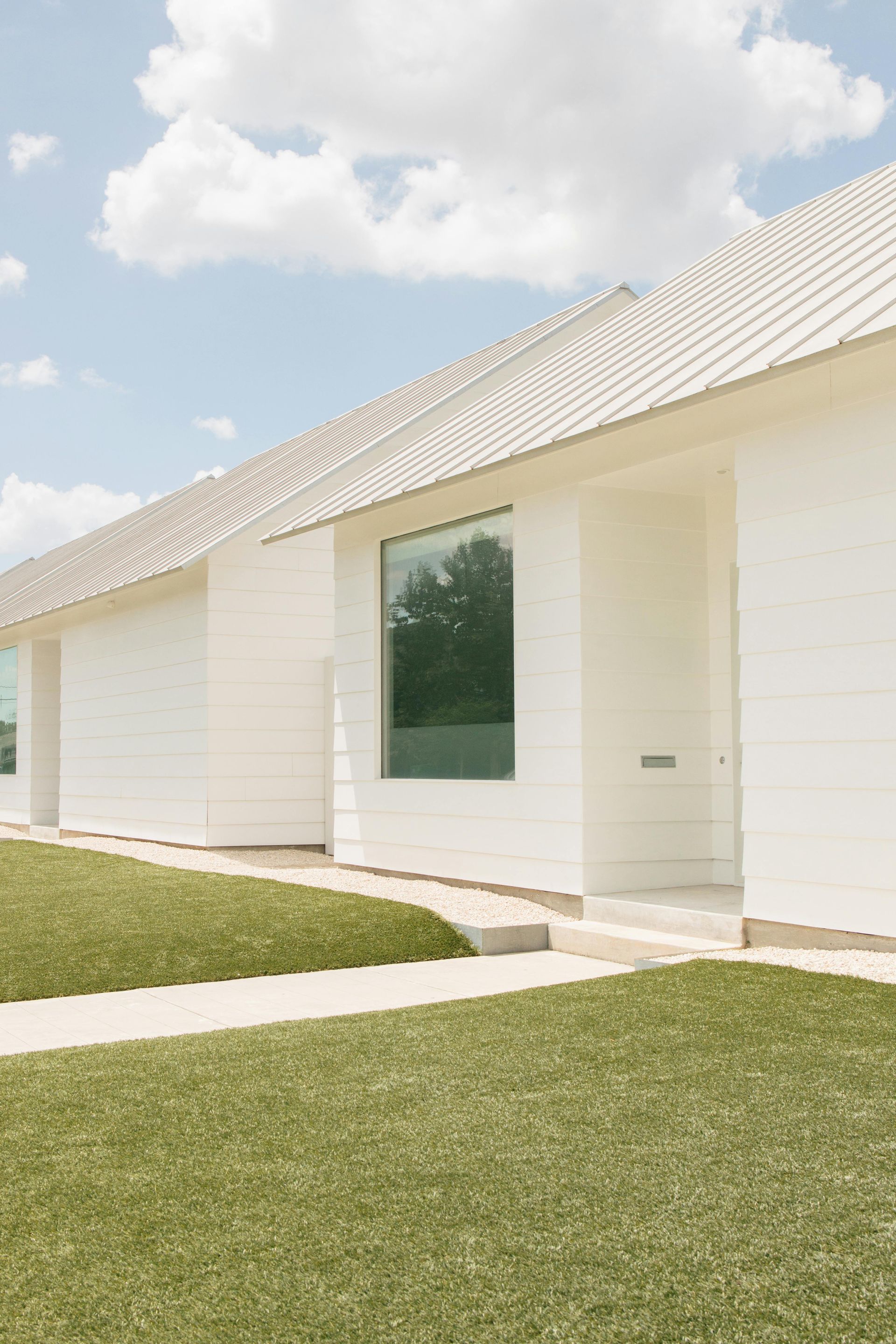White modern home with a green lawn and a large rectangular window reflecting trees, on a sunny day.