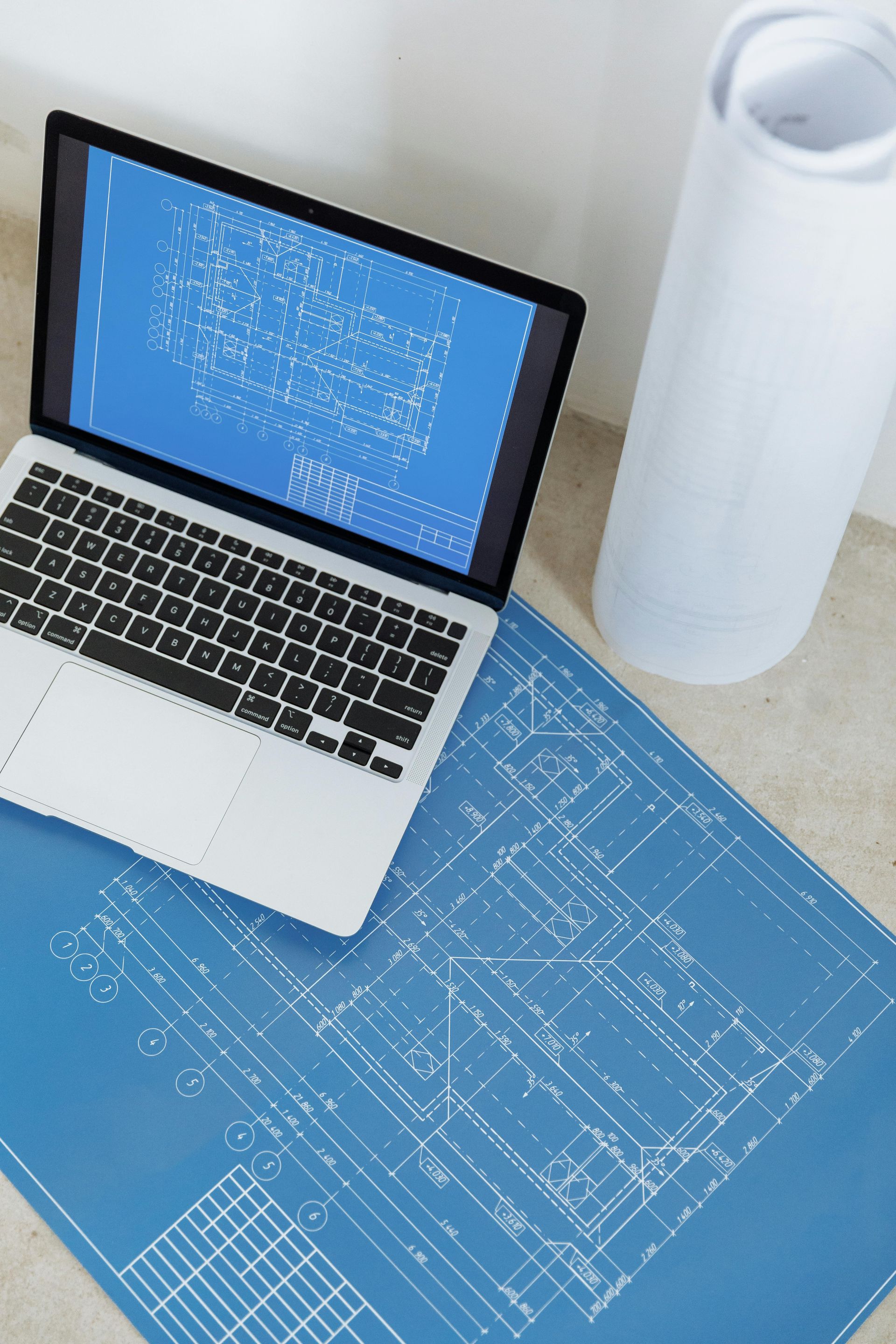 Laptop with blueprint on screen, over a large blueprint, next to rolled-up paper.