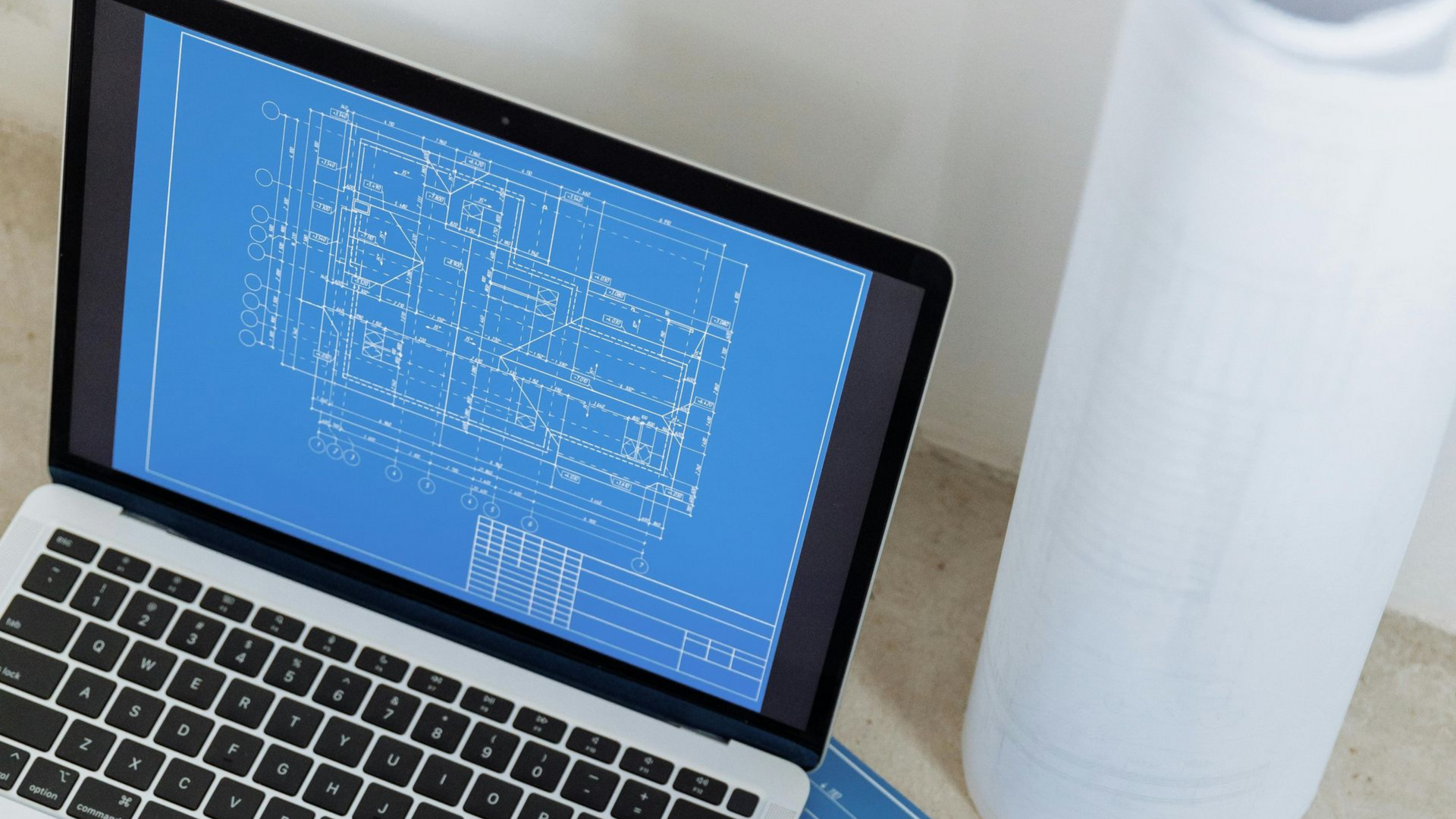 A laptop open on a table displays a blue architectural floor plan next to a white cylindrical object.