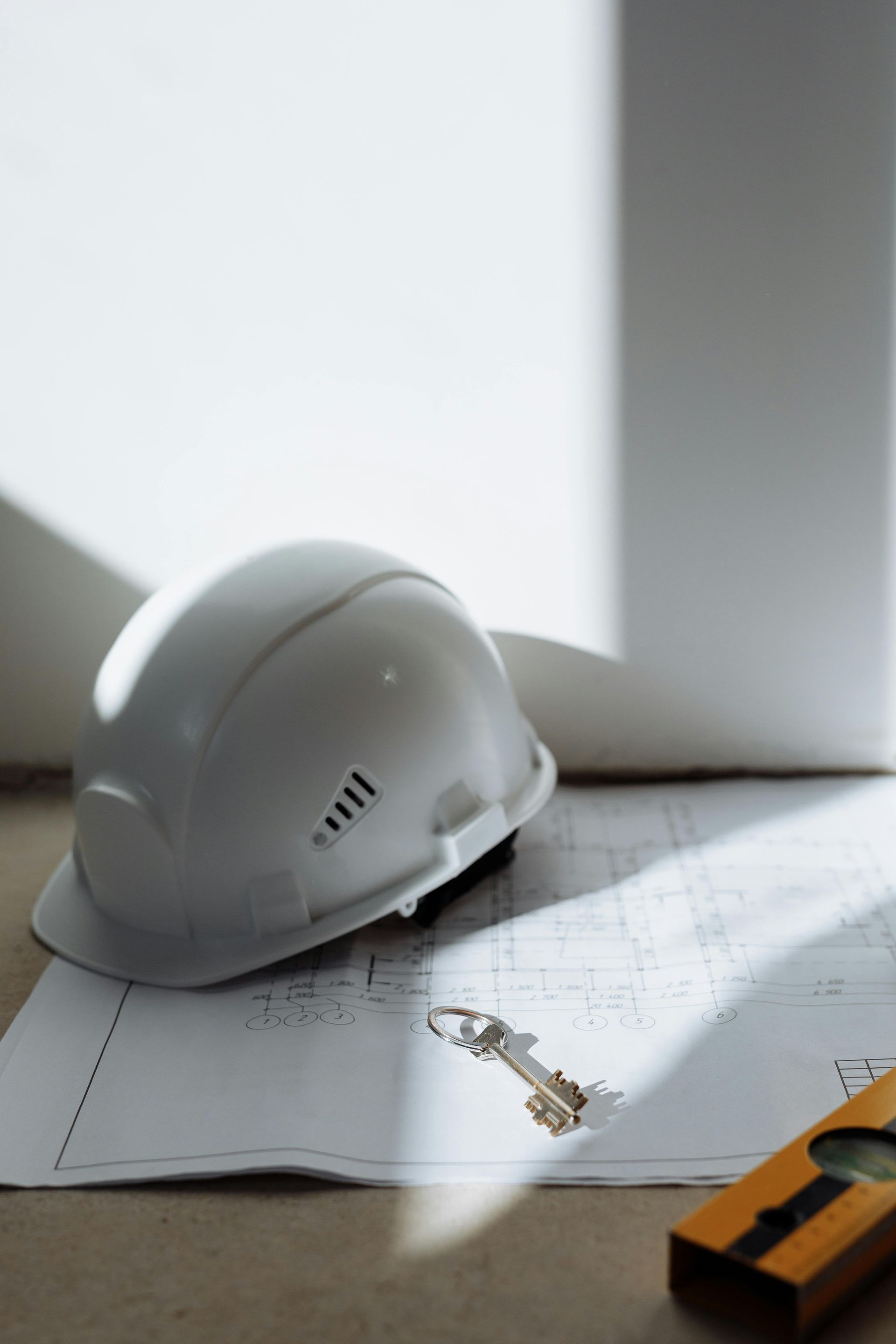 White hard hat, blueprint, key, and level on a white wall and floor.
