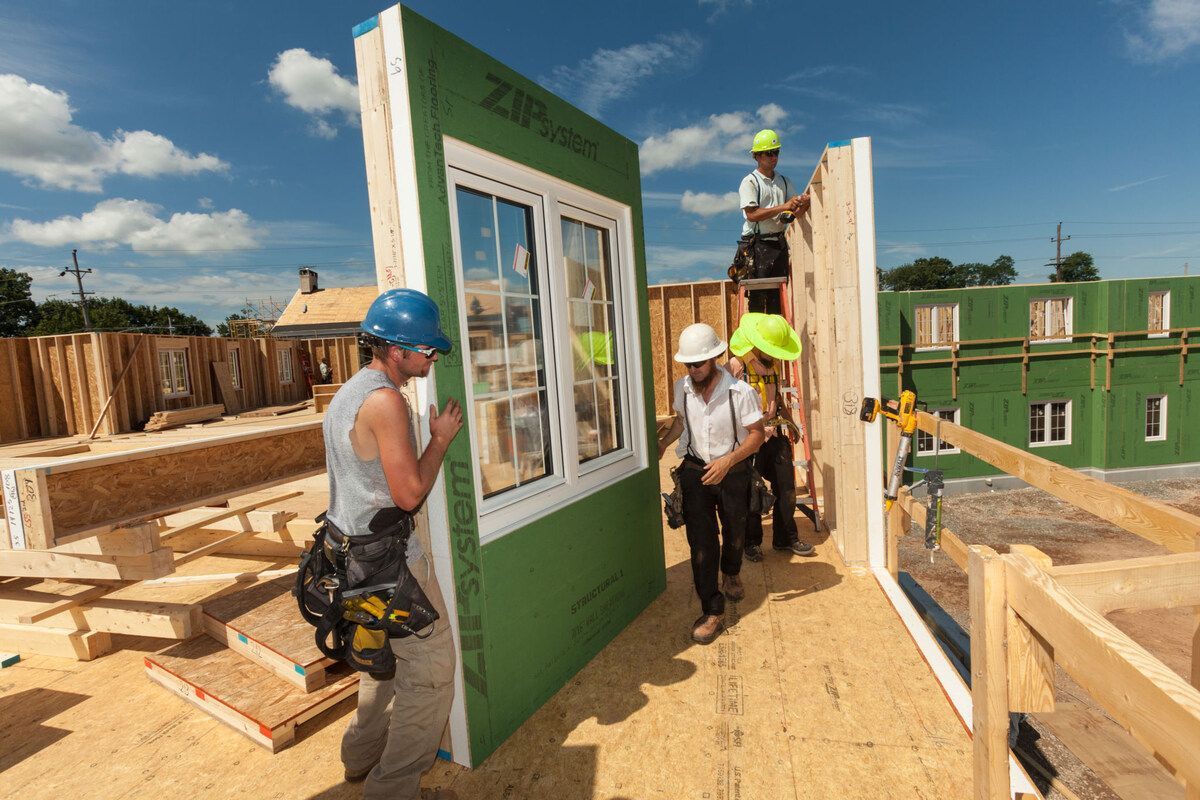 Construction workers in safety gear install a green panel wall with a pre-installed window on a sunny building site.