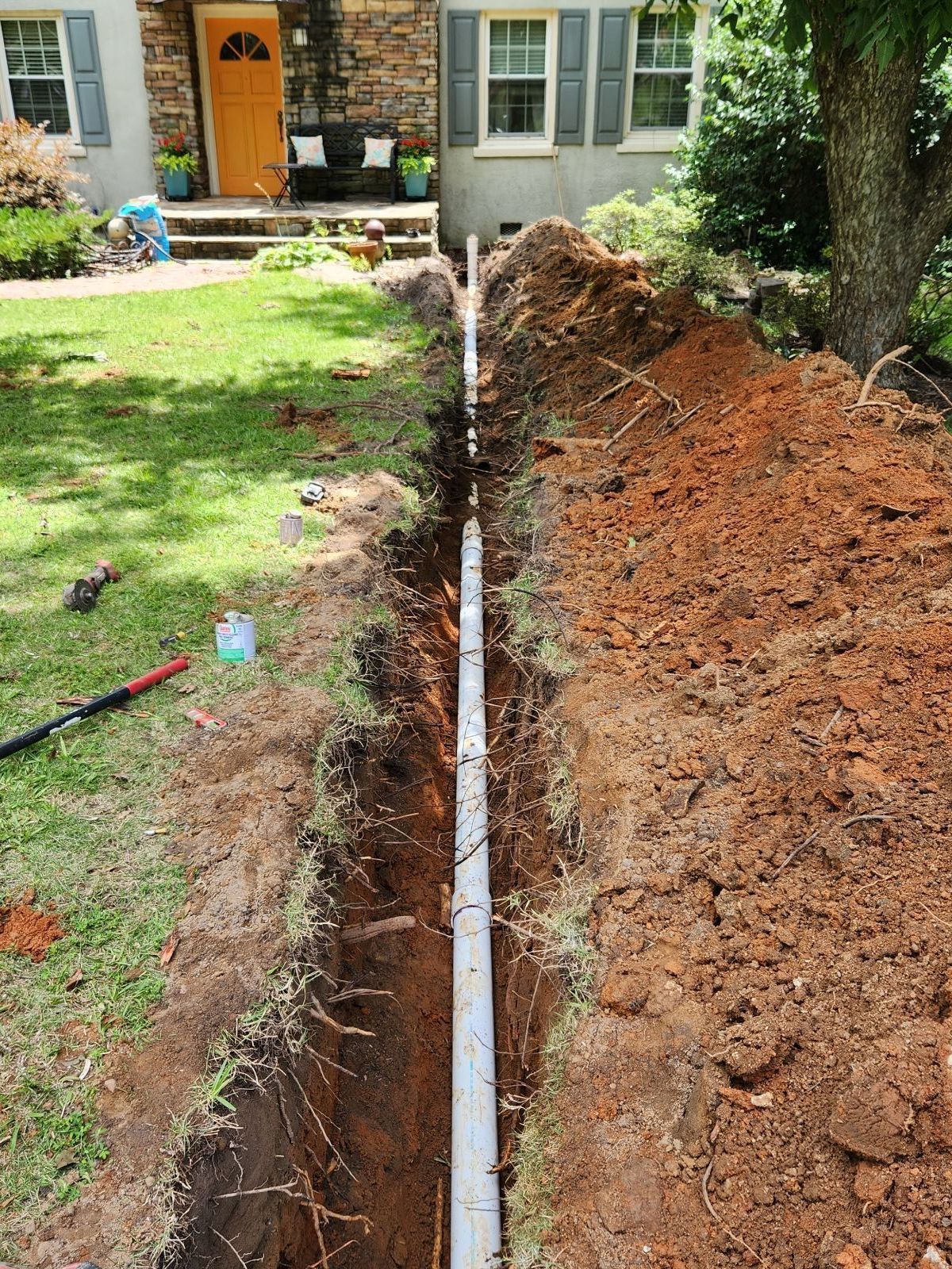 A trench dug in front of a house with a pipe installed. The ground is dirt and grass.