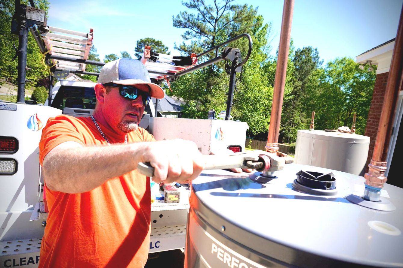 A plumber in orange shirt working on a water heater outside, holding a wrench.