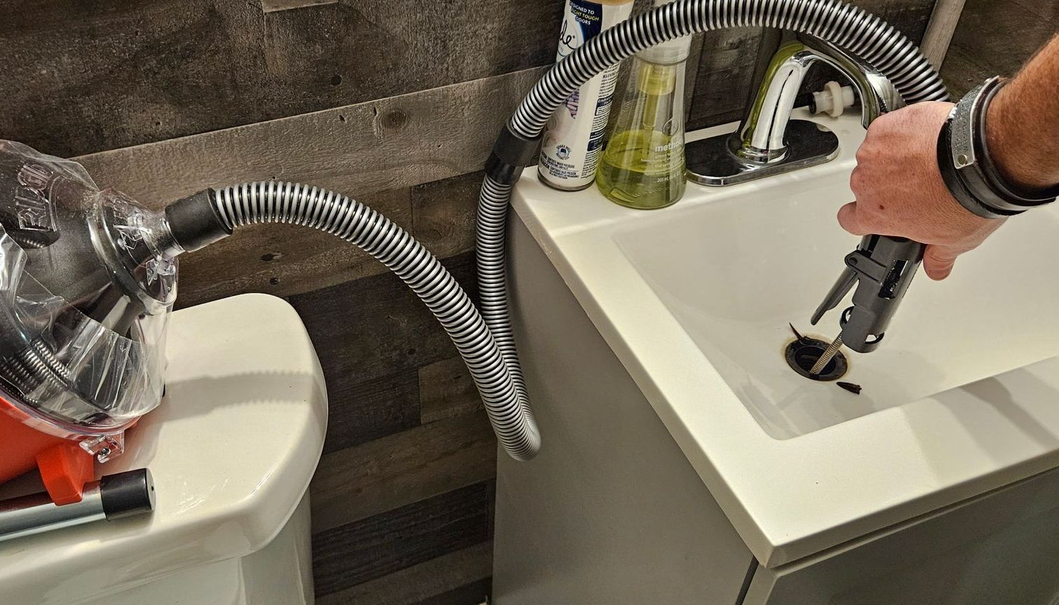 Person using a vacuum hose to try and unclog a bathroom sink, with toilet visible.