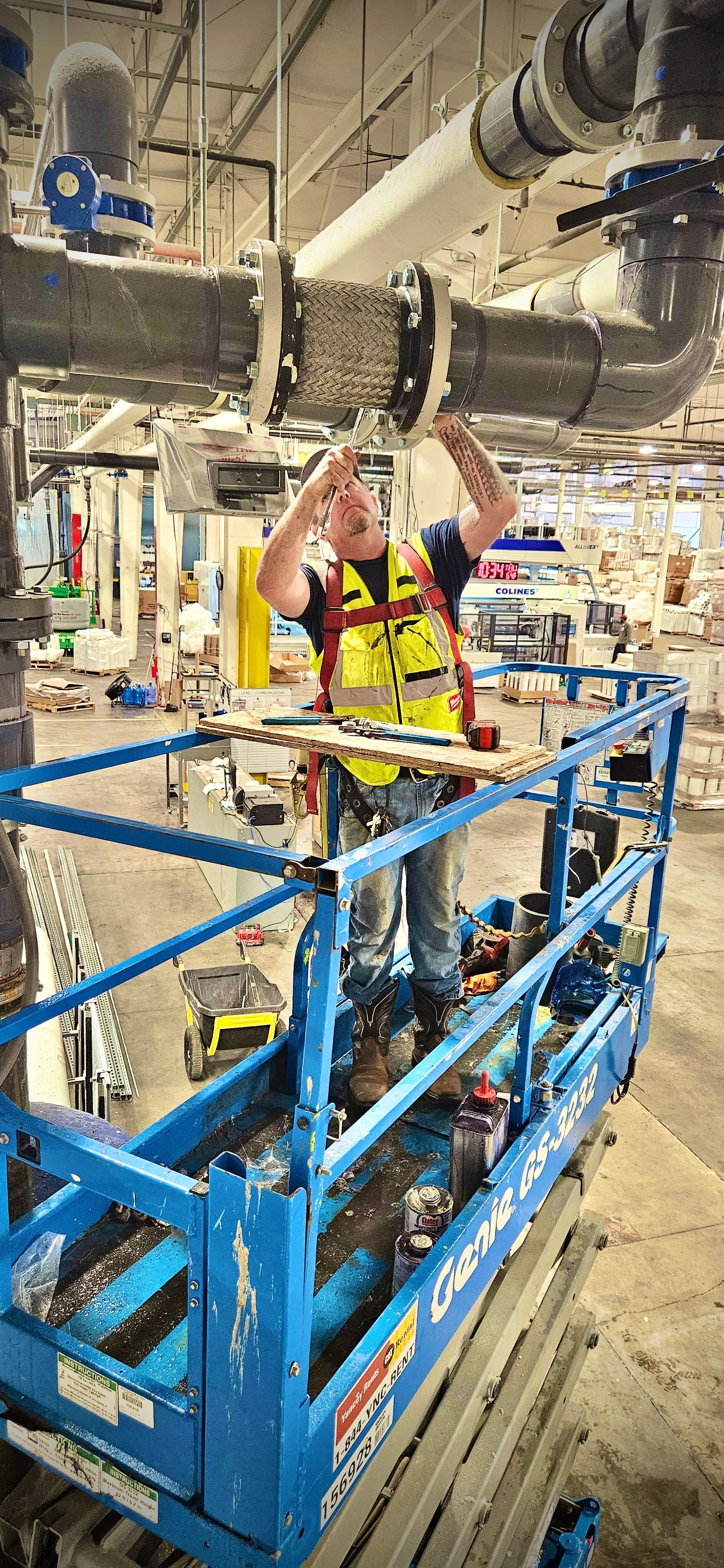 A worker on a blue lift repairs overhead pipes in a large industrial setting. He wears a safety vest.