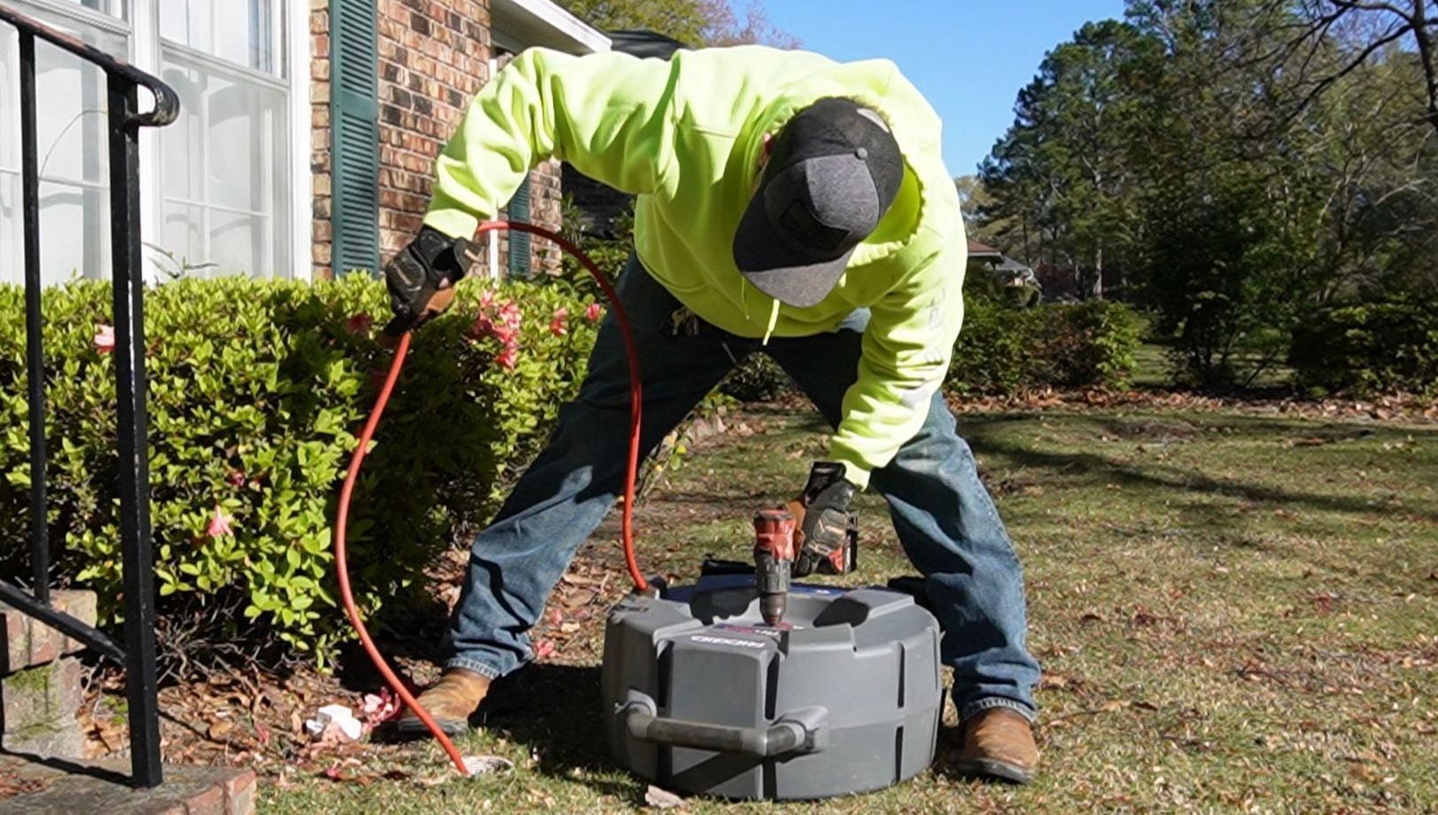 Man in safety gear using drain snake to clear a pipe near a house.