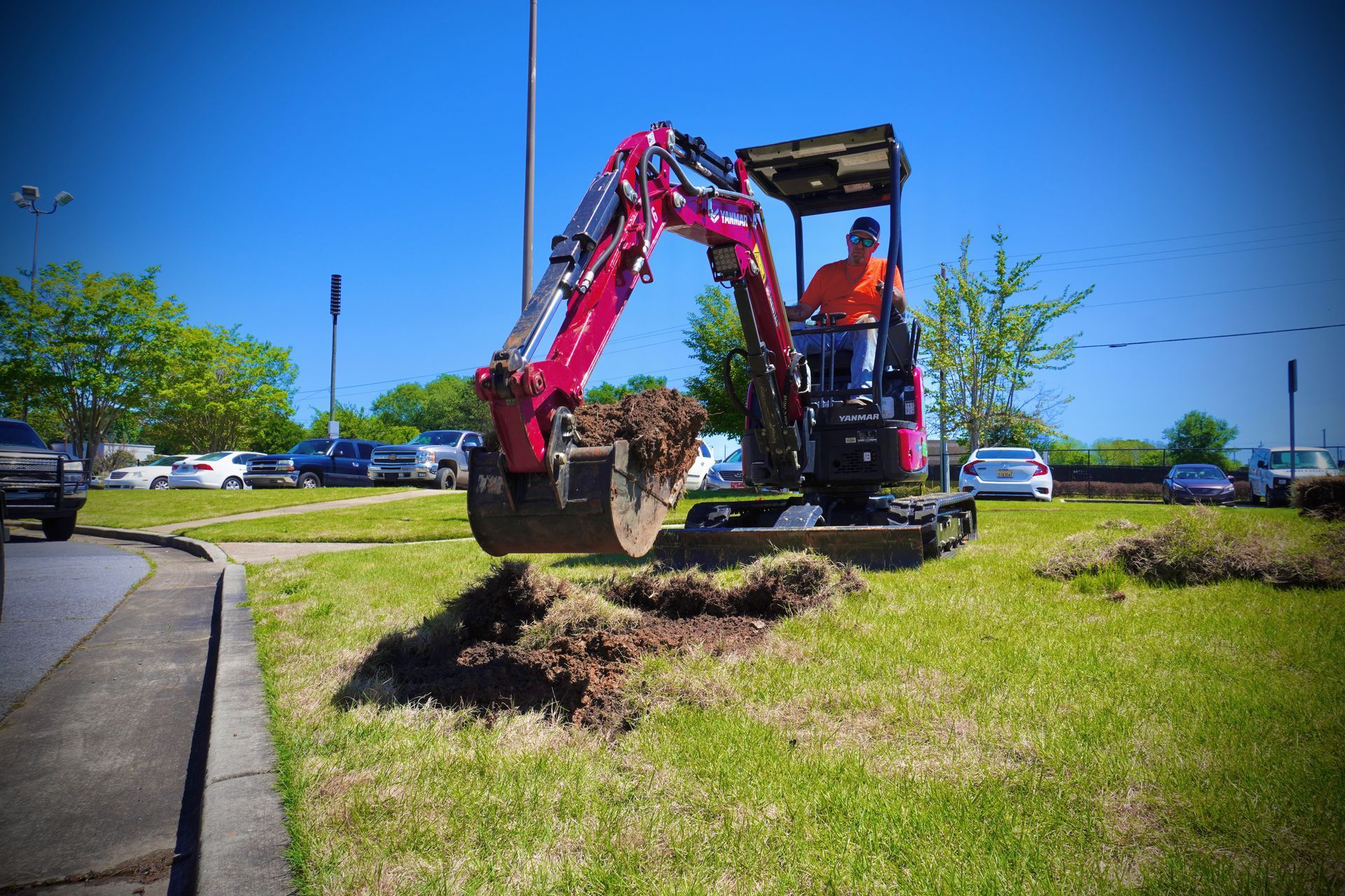 A burgundy mini excavator digging in a grassy area with a person operating it on a sunny day.