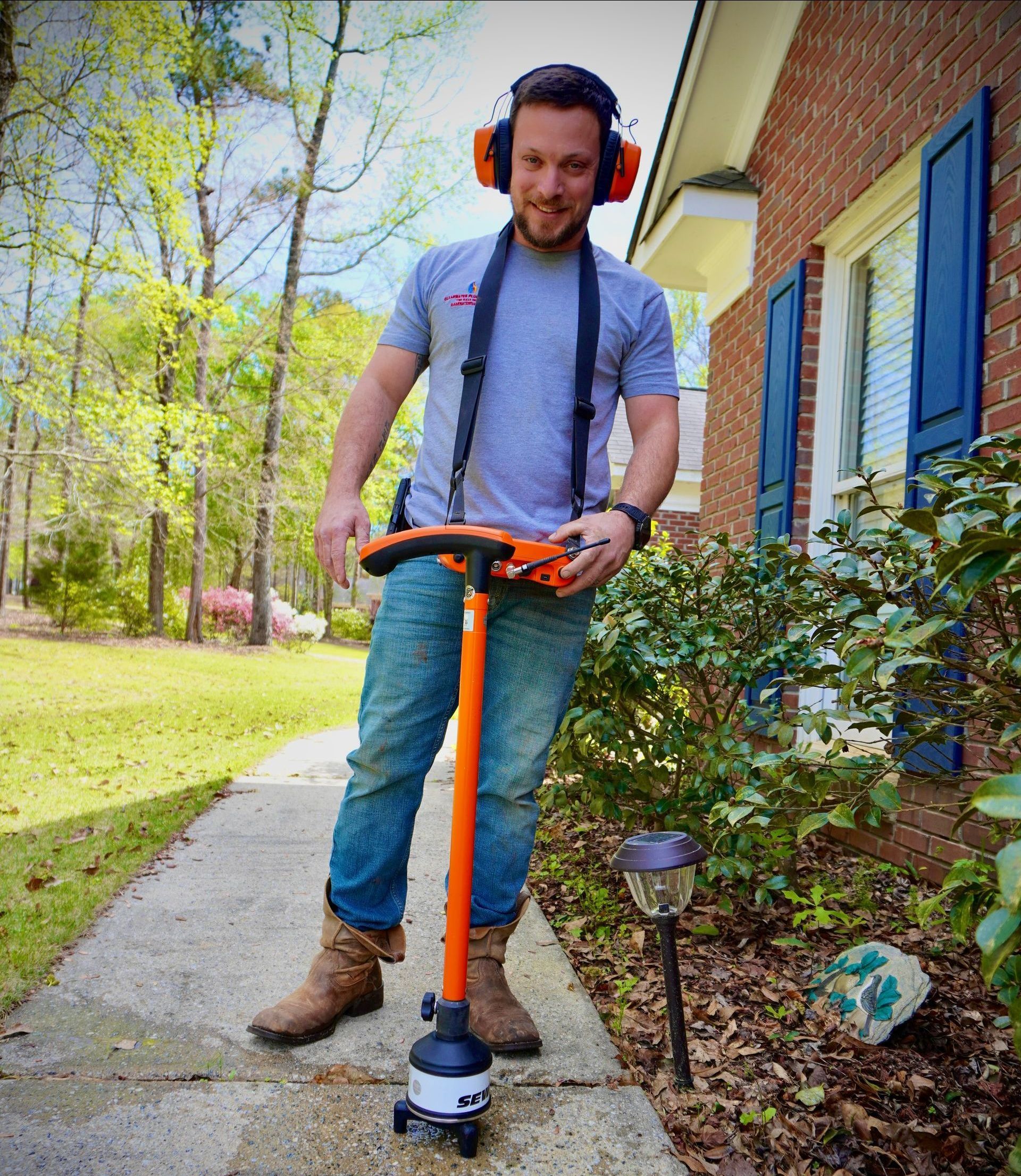 Man using ground penetrating radar device on a sidewalk near a house, wearing ear protection.