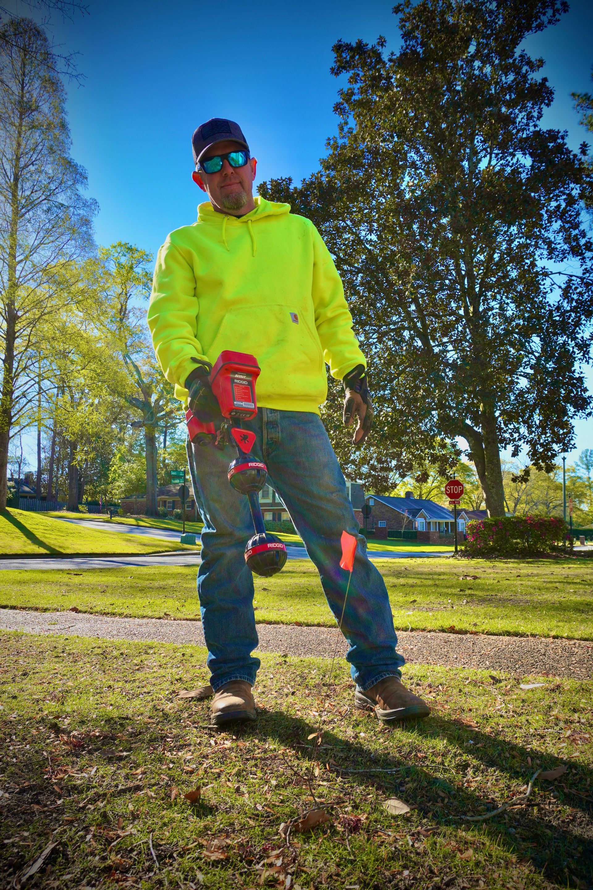 Man in neon hoodie, jeans, and sunglasses holds a red tool outdoors; park setting.