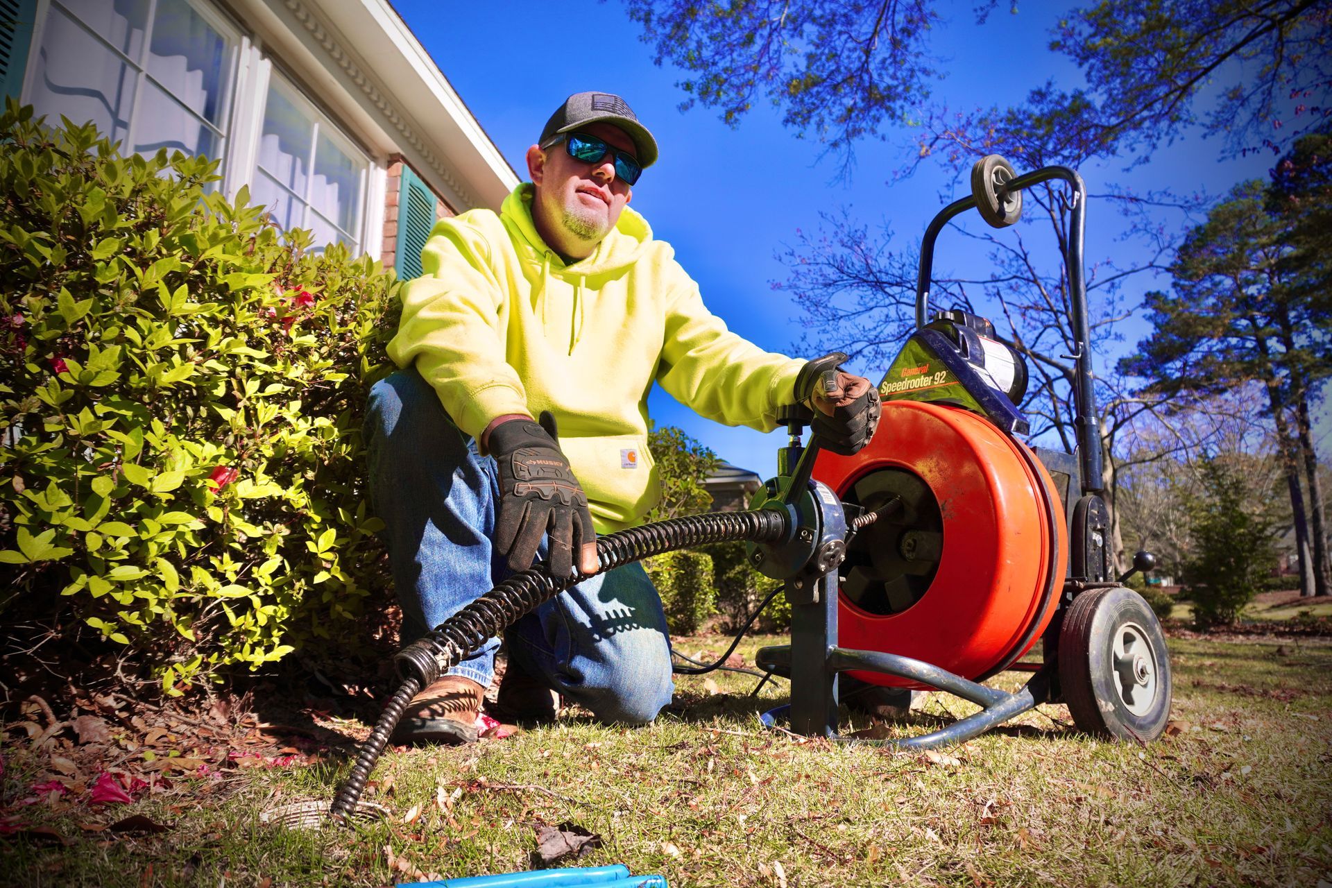 Plumber kneeling outside, operating a drain cleaning machine. Bright yellow sweatshirt, blue jeans, sunny day.