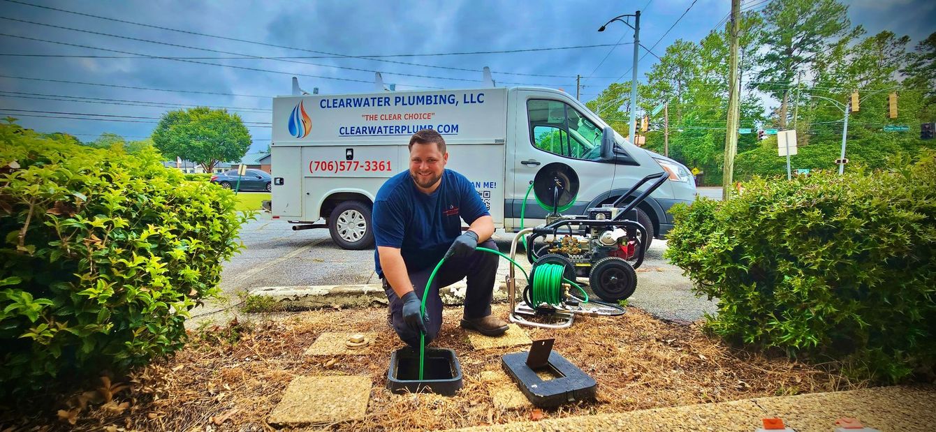 A plumber kneels near a drain, preparing to use equipment. A van with the company logo is behind him.