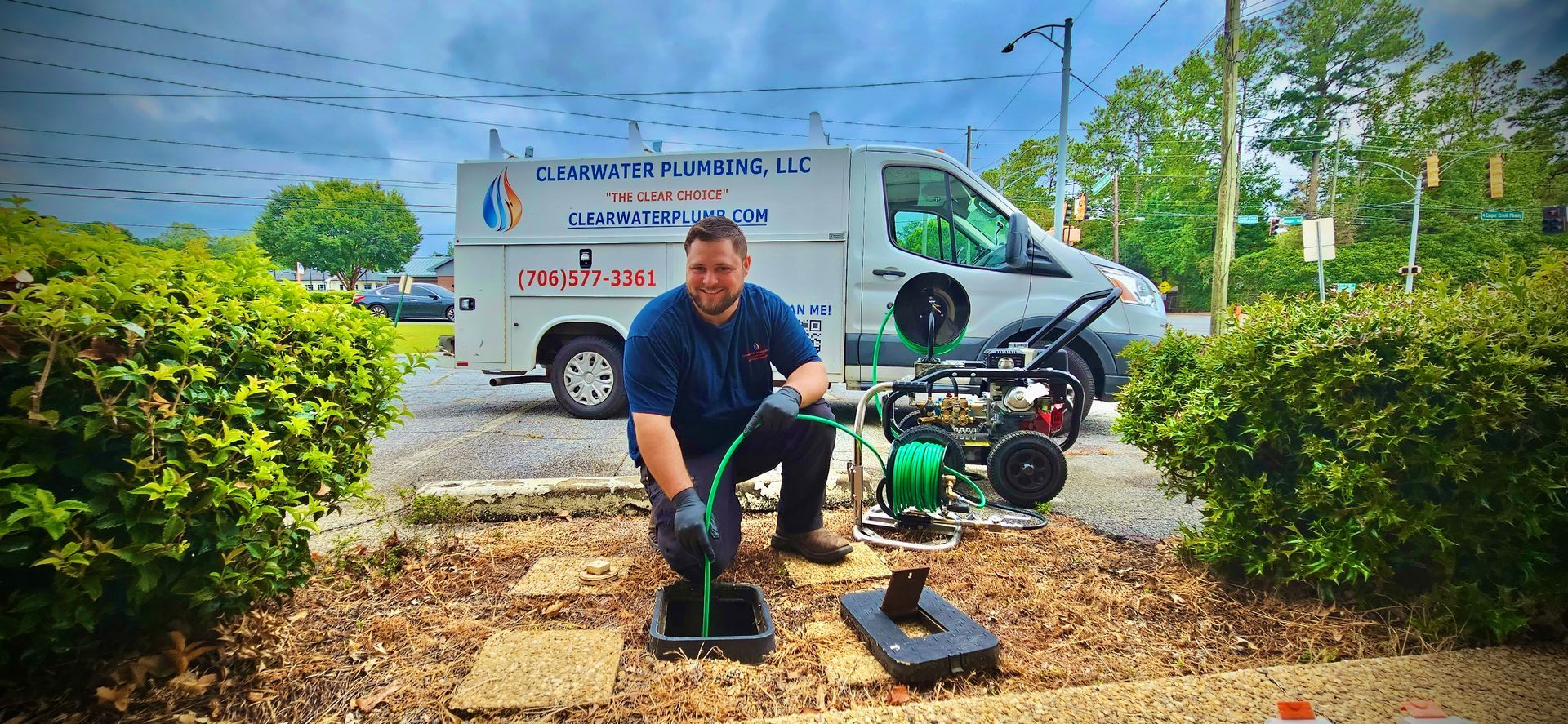 Plumber kneeling, working on a drain; van and equipment in background, shrubs, cloudy sky.