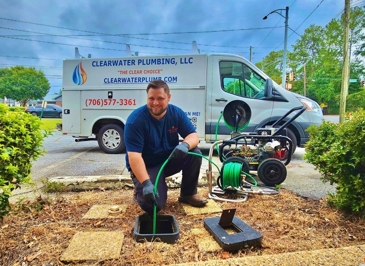 Plumber kneeling, unclogging drain with a hose, in front of a service van; brown yard.
