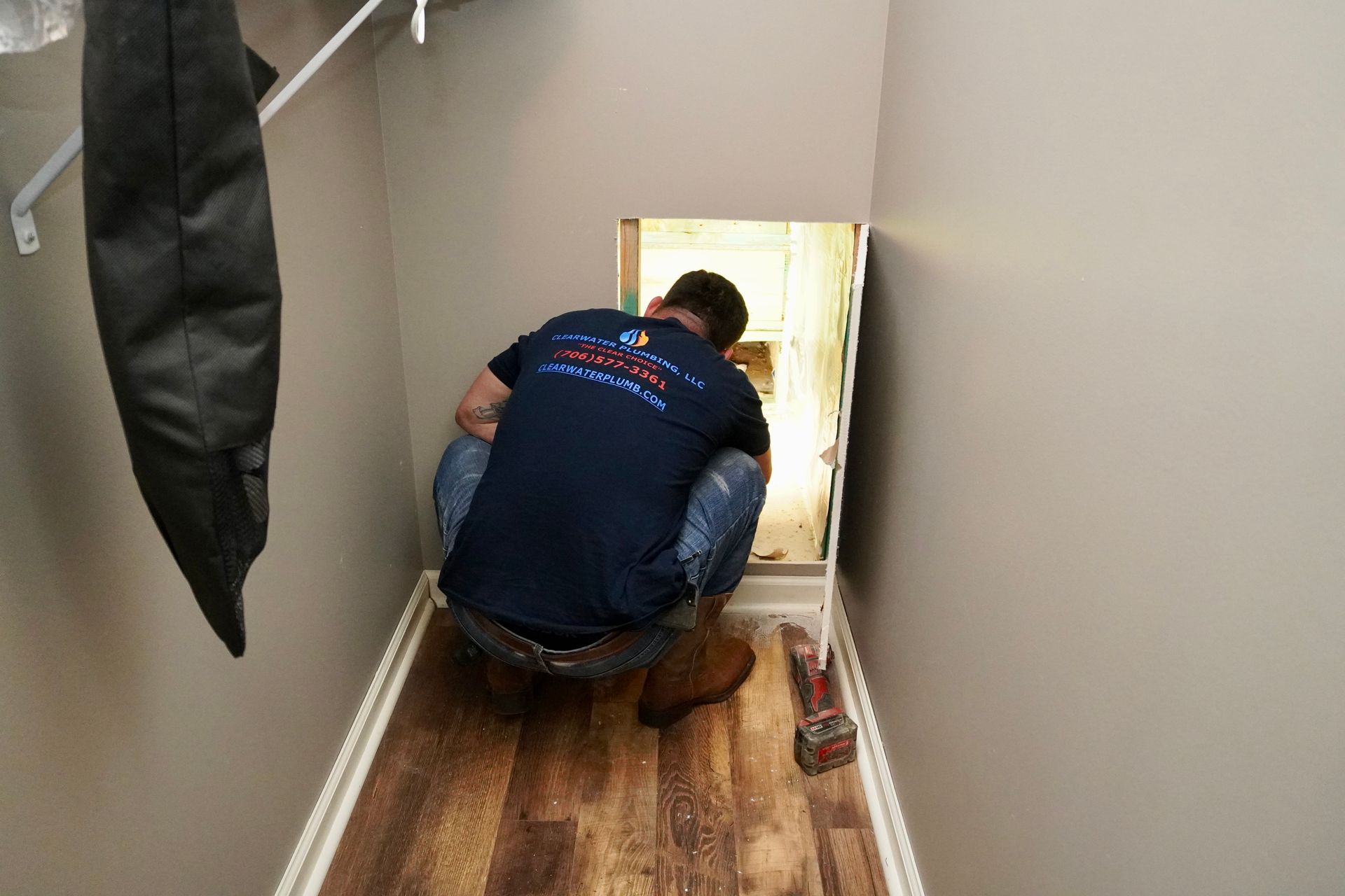 Man crouches in a narrow crawl space, working. Interior with wood flooring and light gray walls.