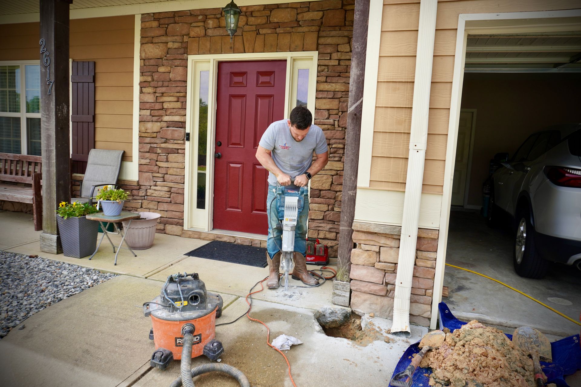 A man uses a jackhammer on concrete outside a home. A wet/dry vacuum and debris are visible.