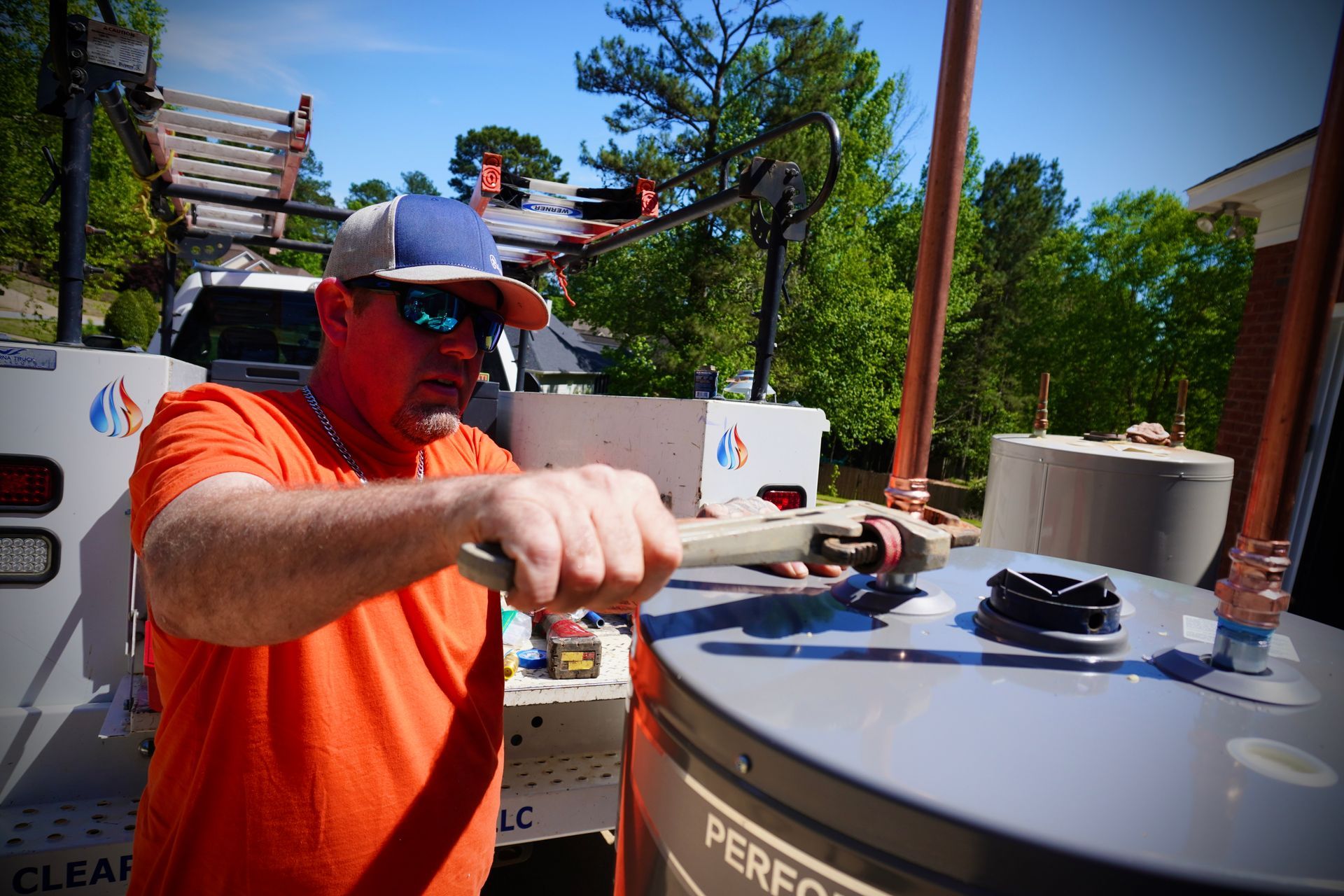 Plumber in an orange shirt works on a water heater outdoors, holding a wrench.