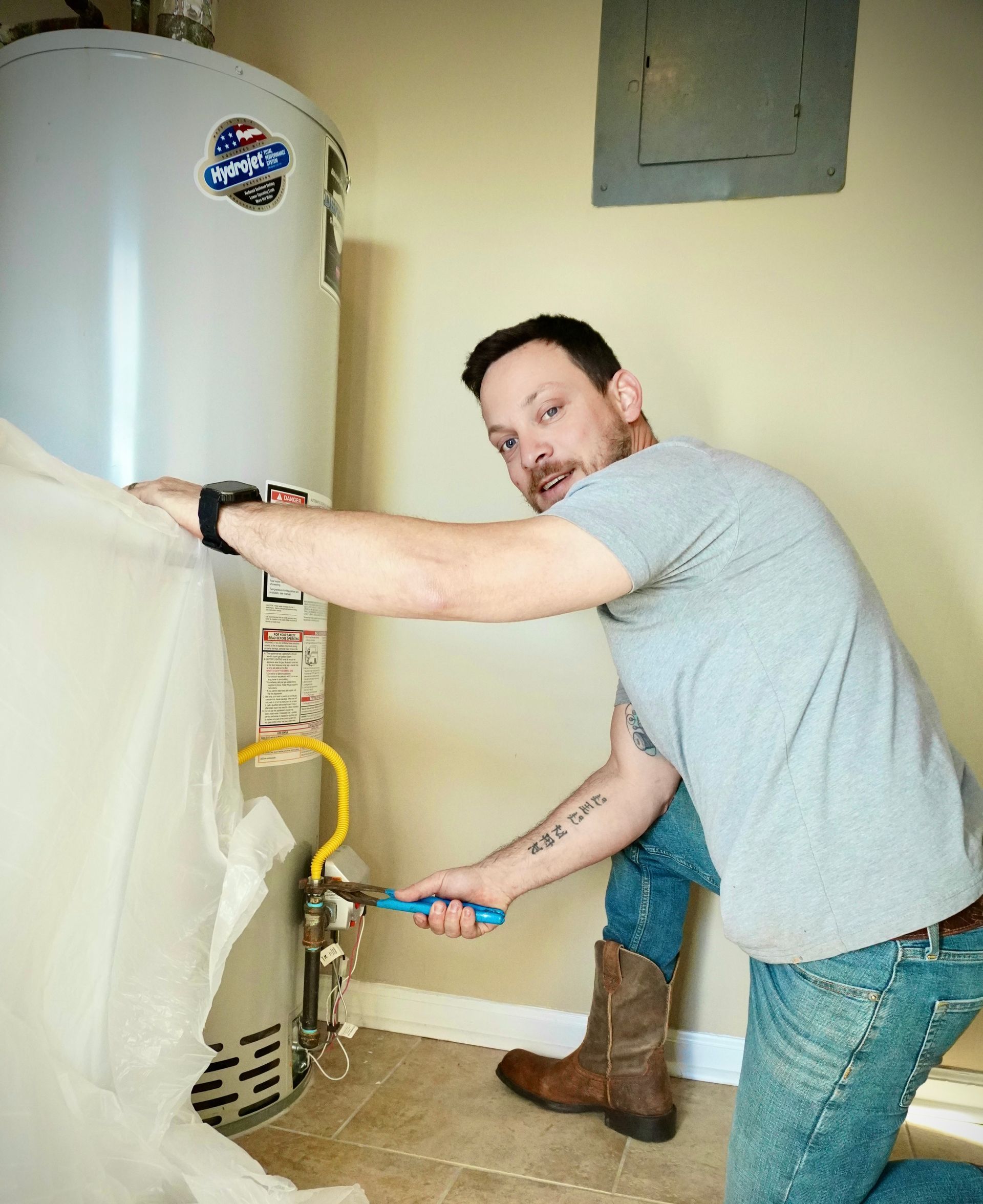 Man in jeans kneels, working on a water heater with a wrench. Indoor setting, beige walls, brown boots.