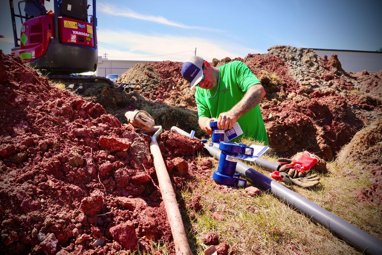 Man in green shirt and cap working on pipes in a dirt trench; excavator in background.
