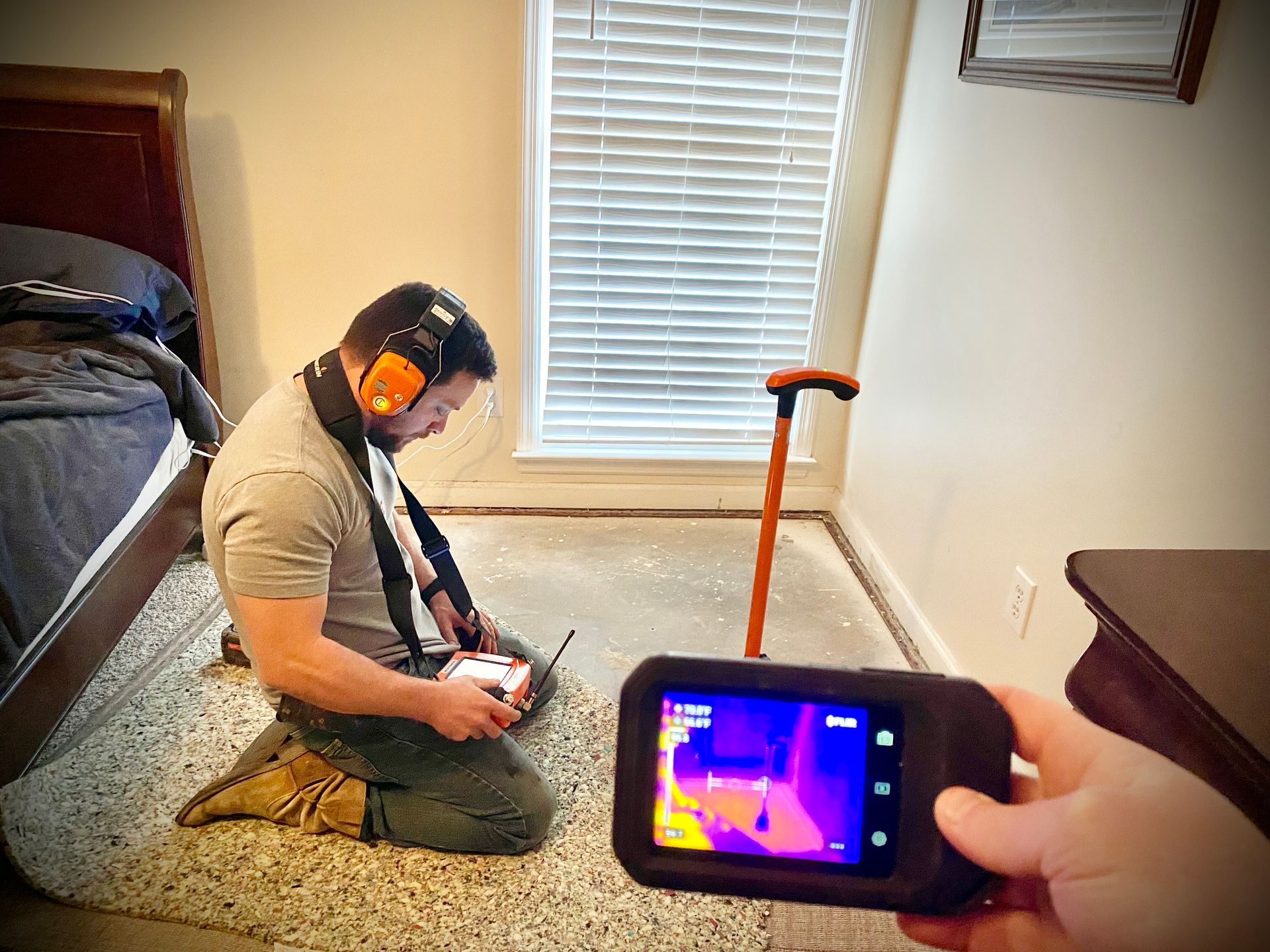 Man uses a thermal camera to inspect a room's floor near a window. The camera displays heat signatures in colorful tones.