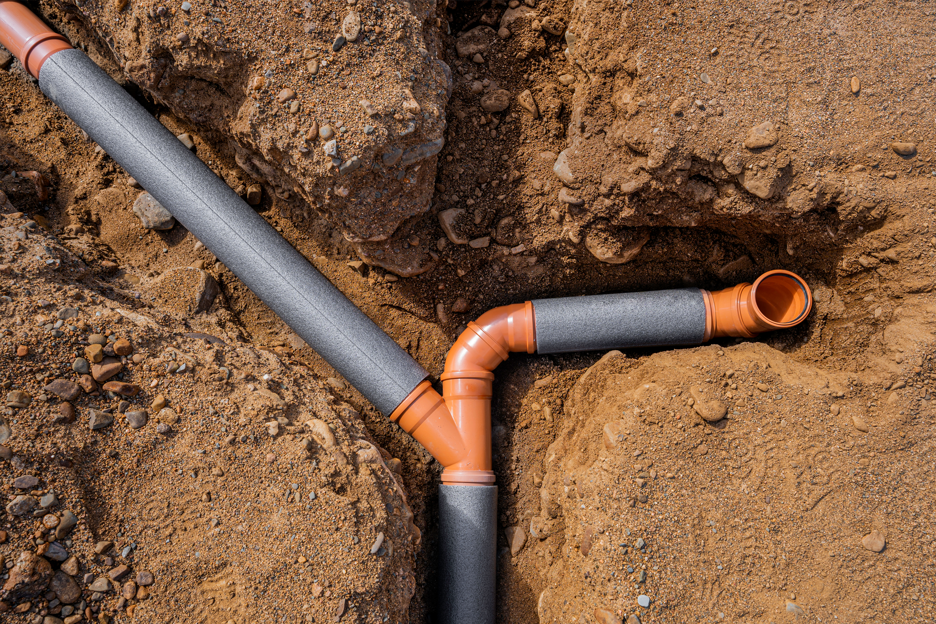 Orange plastic drain pipes installed in a trench, some wrapped in grey insulation, in brown dirt.