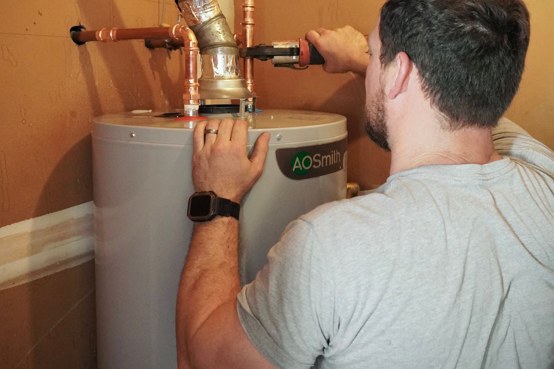 Man tightening pipe fitting on top of a water heater in a utility room.