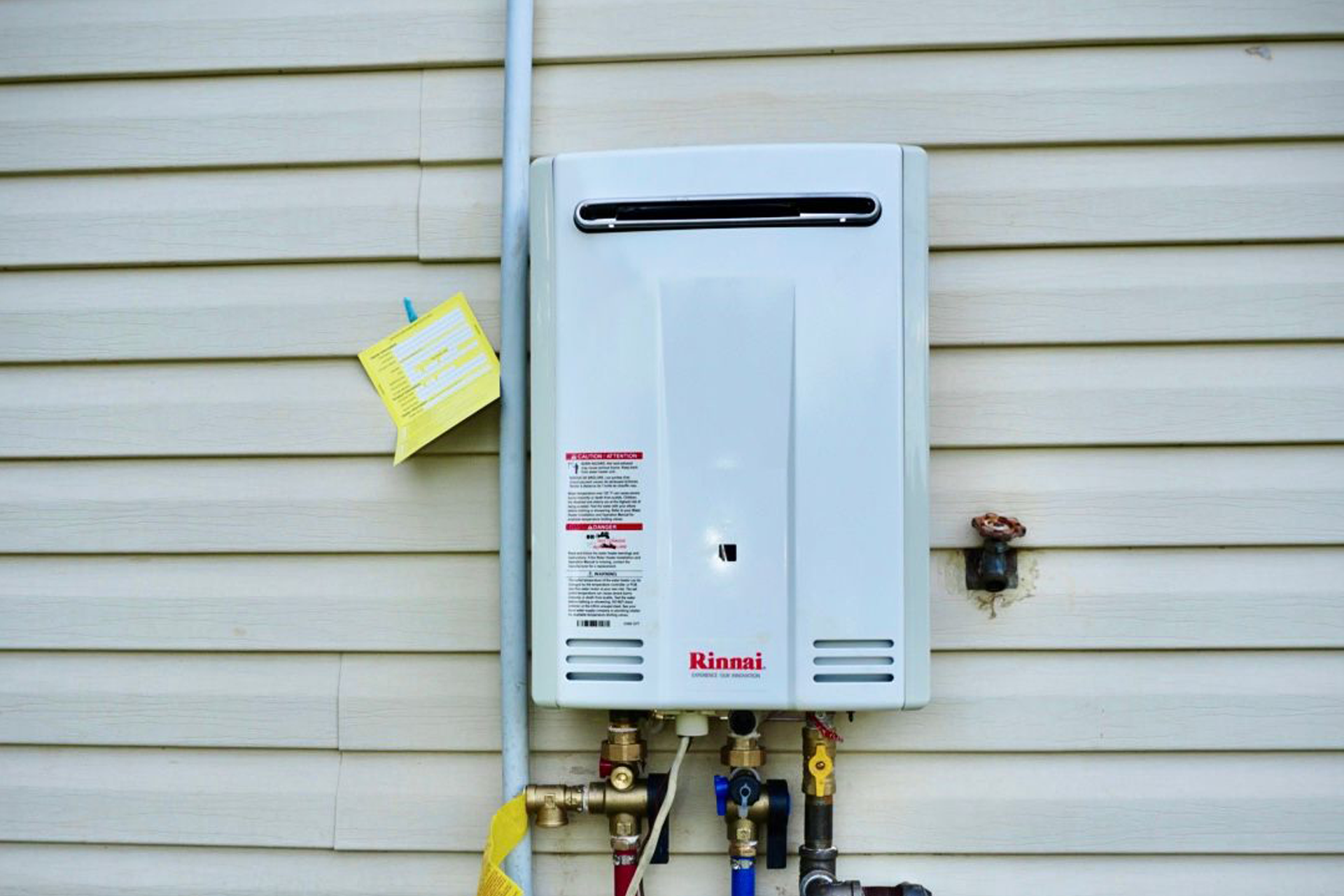 White tankless water heater on beige siding, with pipes and a yellow tag.