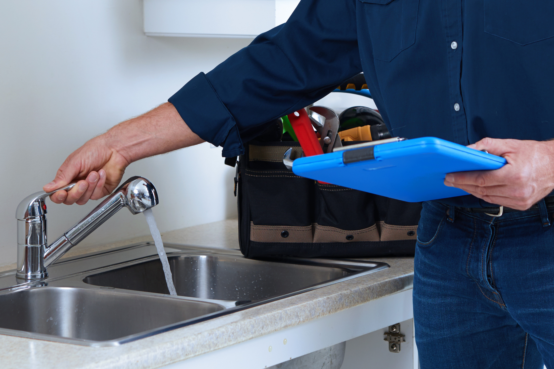 Plumber turning on kitchen faucet, checking water flow. Holding clipboard, toolbox in background.