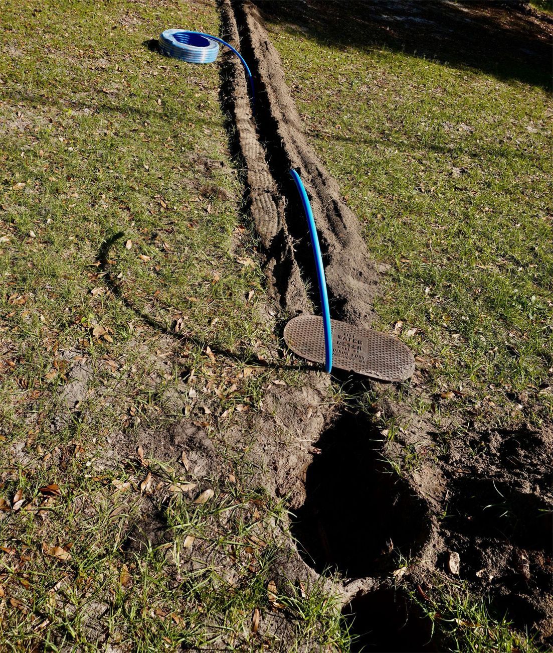 A long trench in a grassy yard, with blue pipe laid inside and a dark circular hole.