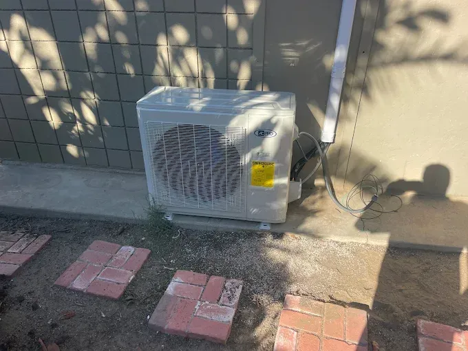 White air conditioner unit against a textured wall, yellow sticker visible. Red bricks border ground.