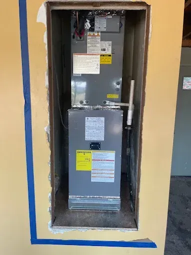 Furnace unit installed in a wall opening, inside a house. Yellow wall with blue tape.