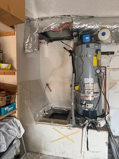 Water heater and access panel in a utility area with ductwork and a storage shelf.