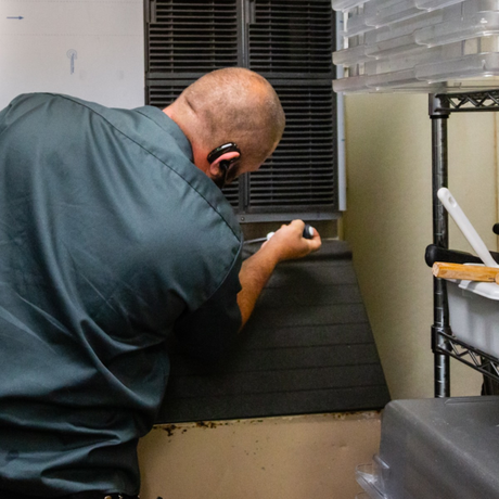 Man in dark shirt leaning over ice bin near a metal rack, possibly cleaning or working in a kitchen.