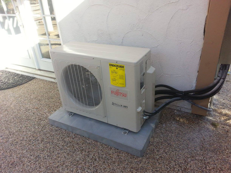 Fujitsu outdoor air conditioning unit on a concrete base, mounted on a gravel surface next to a stucco wall.