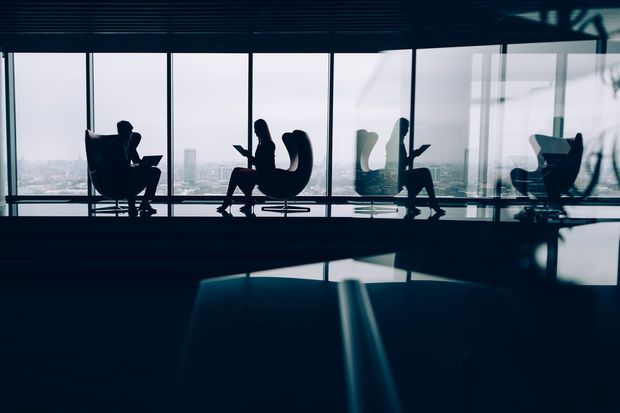 Silhouettes of people sitting in modern armchairs in a dark, high-rise office with large windows overlooking a city.