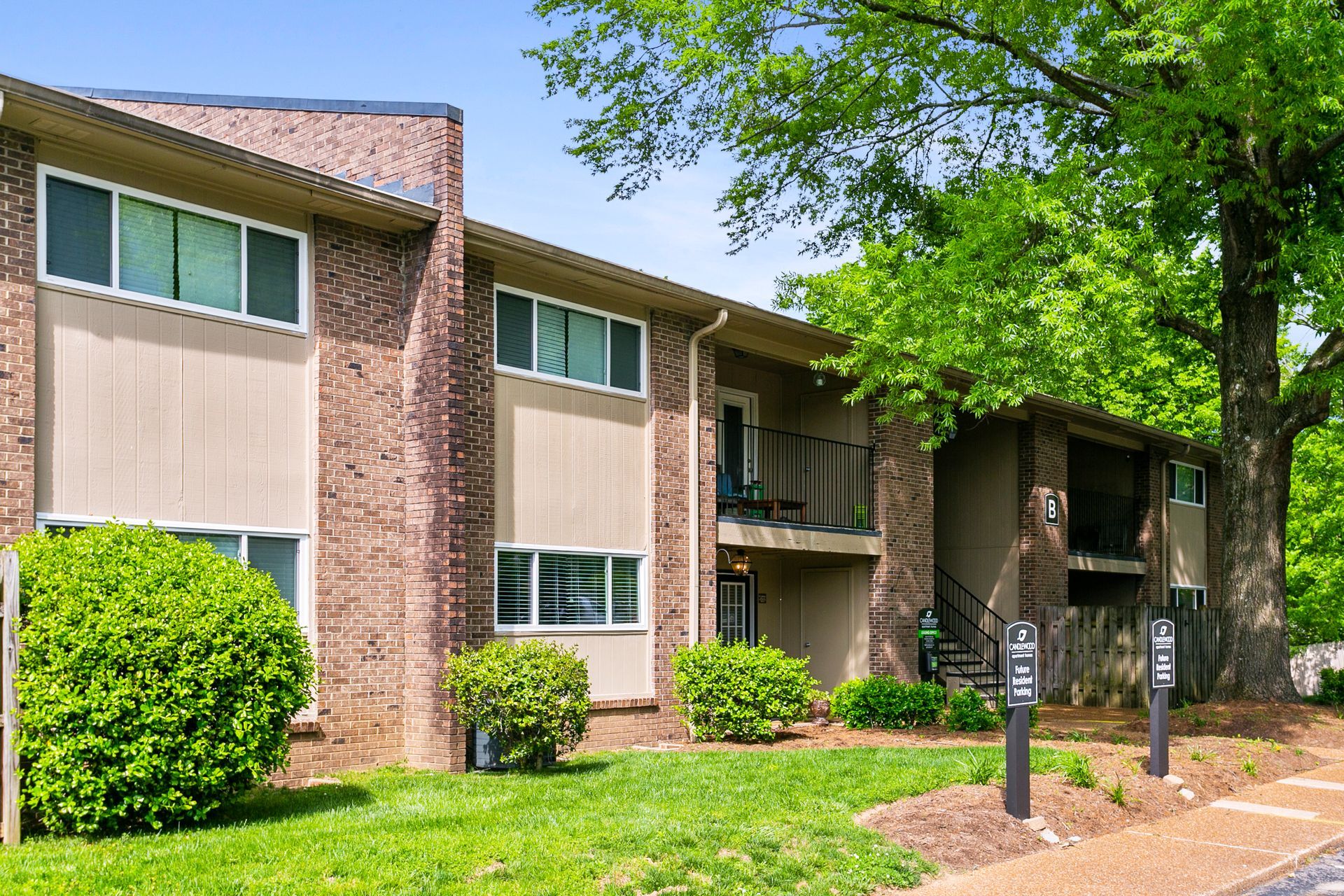 A two-story brick and tan apartment building surrounded by green lawns and trees on a sunny day.