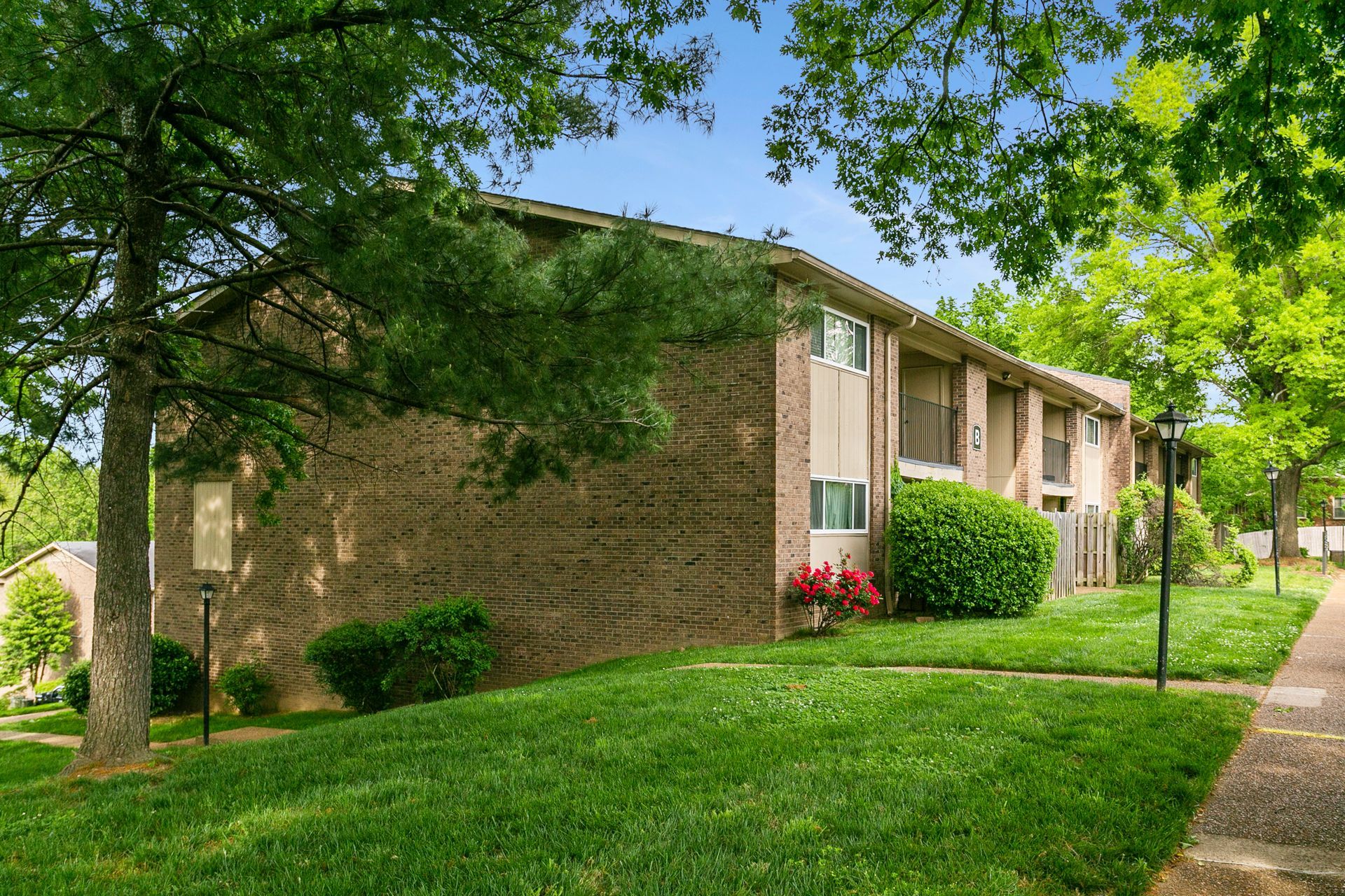 A two-story brick apartment building set on a grassy hill with surrounding trees under a bright blue sky.
