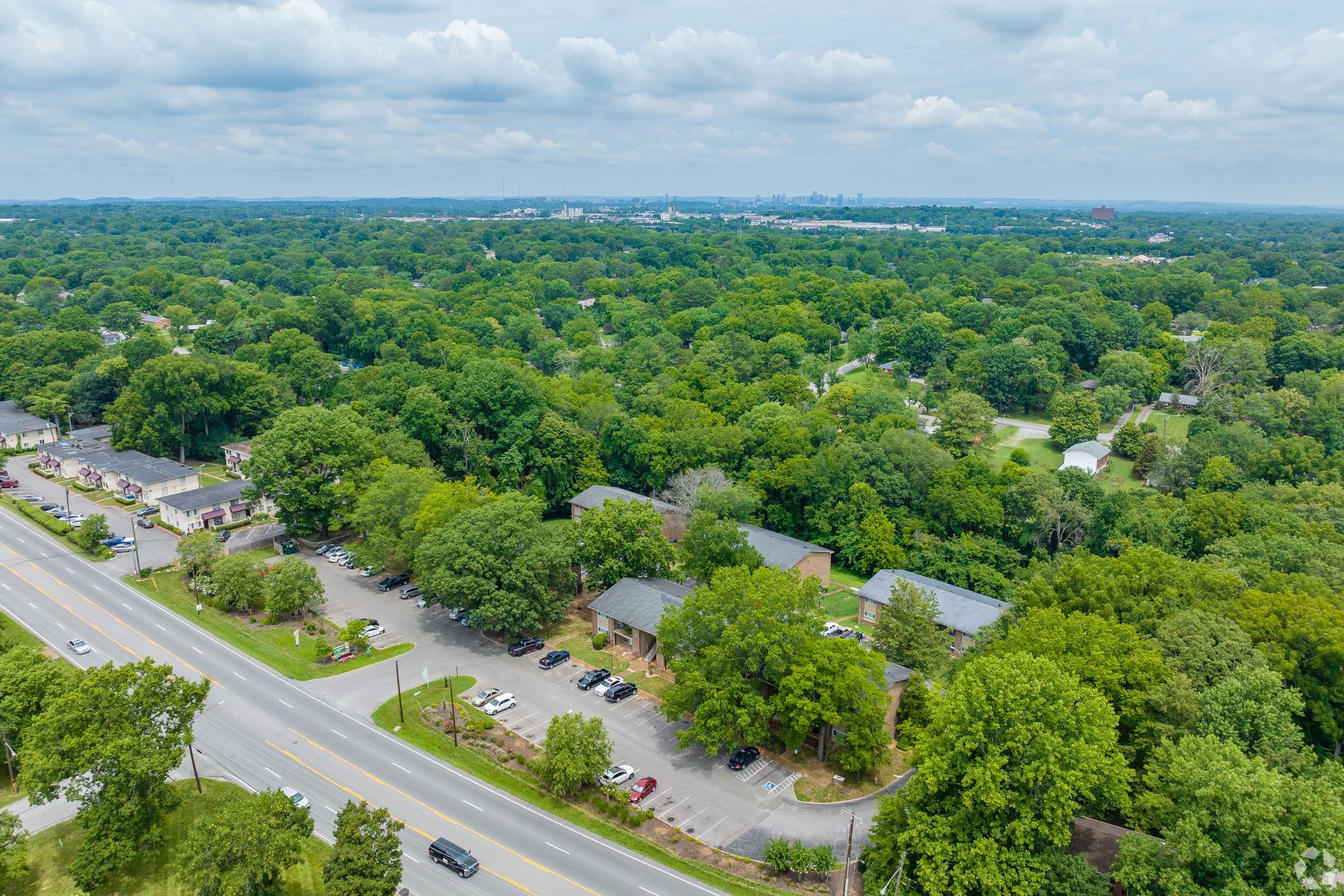 An aerial view of a multi-unit apartment complex nestled among dense green trees beside a road with a parked car.