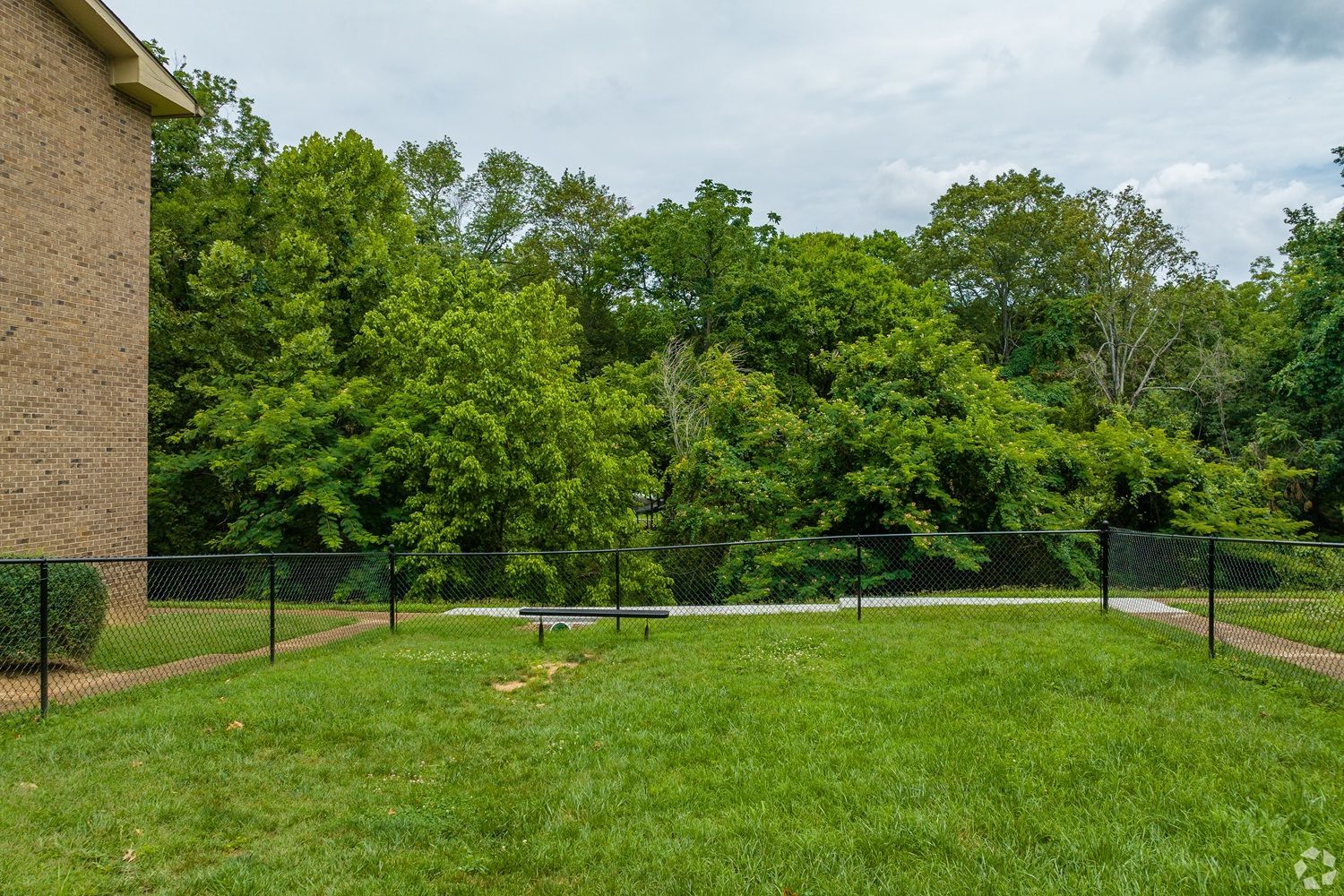 A brick building stands beside a grassy yard enclosed by a chain-link fence, with a dense green forest in the background.