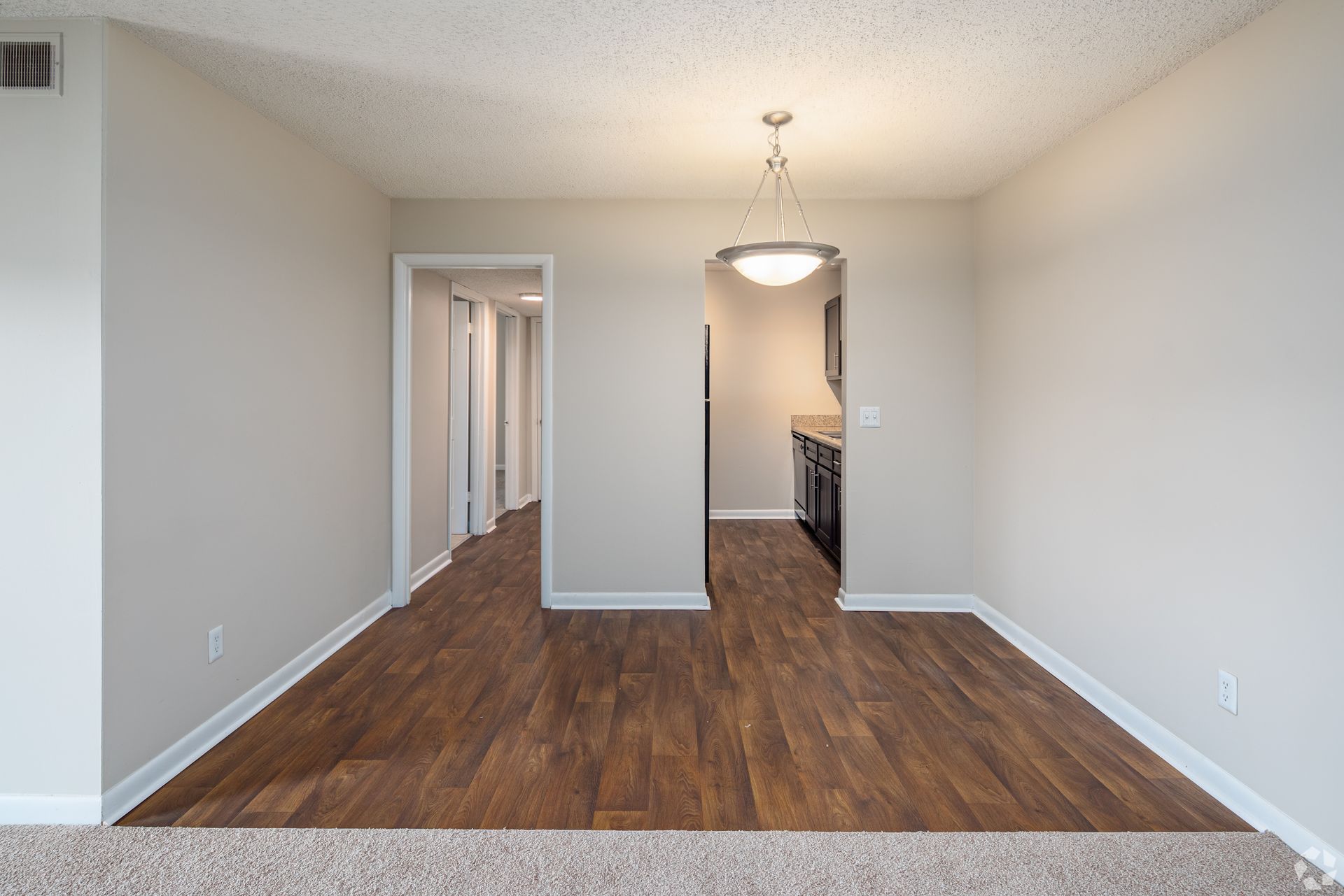 An empty dining area with wood-look flooring, beige walls, a hanging pendant light, and doorways to a kitchen and hallway.