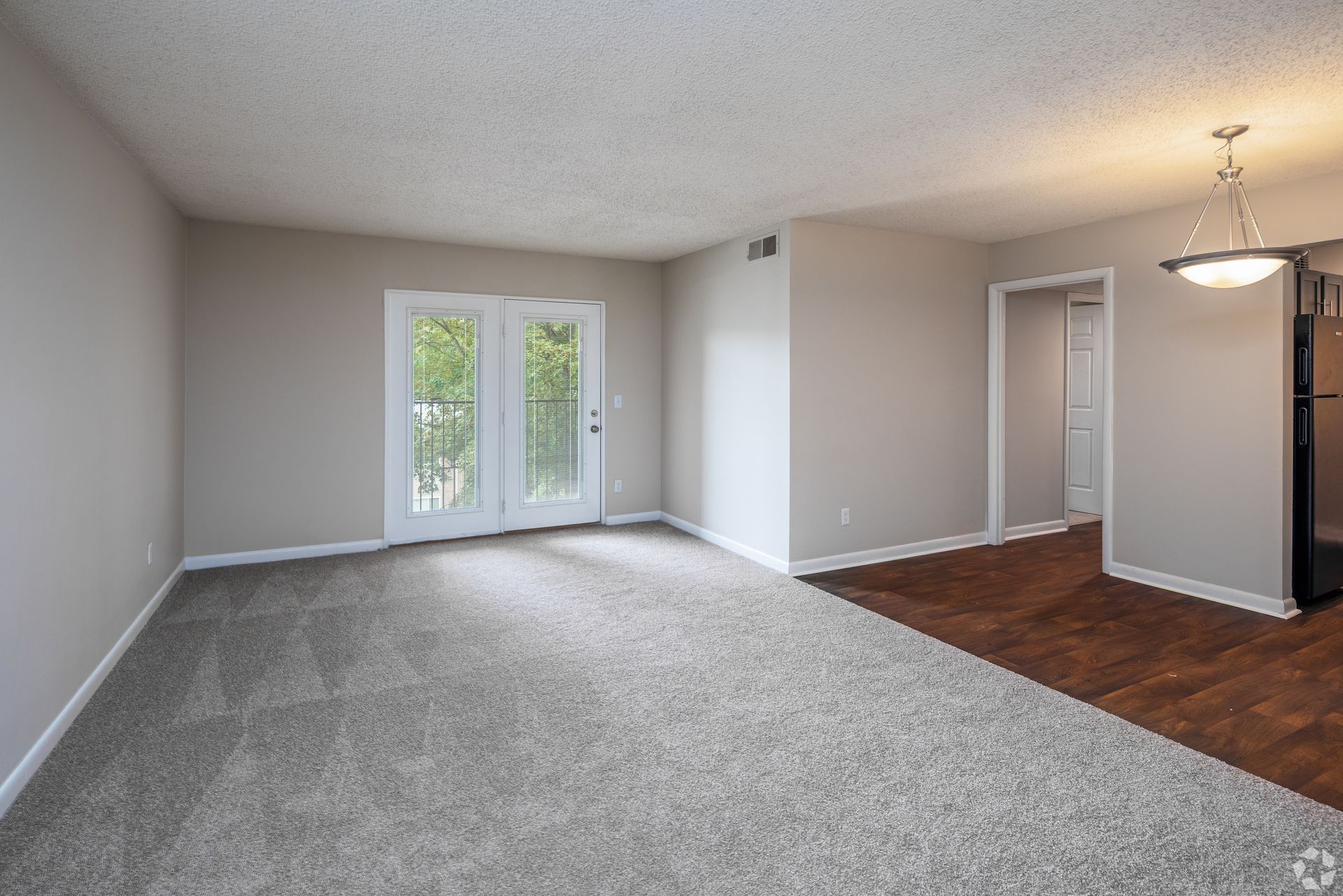 An unfurnished living area with light grey carpet, neutral-colored walls, glass balcony doors, and a dining room pendant.