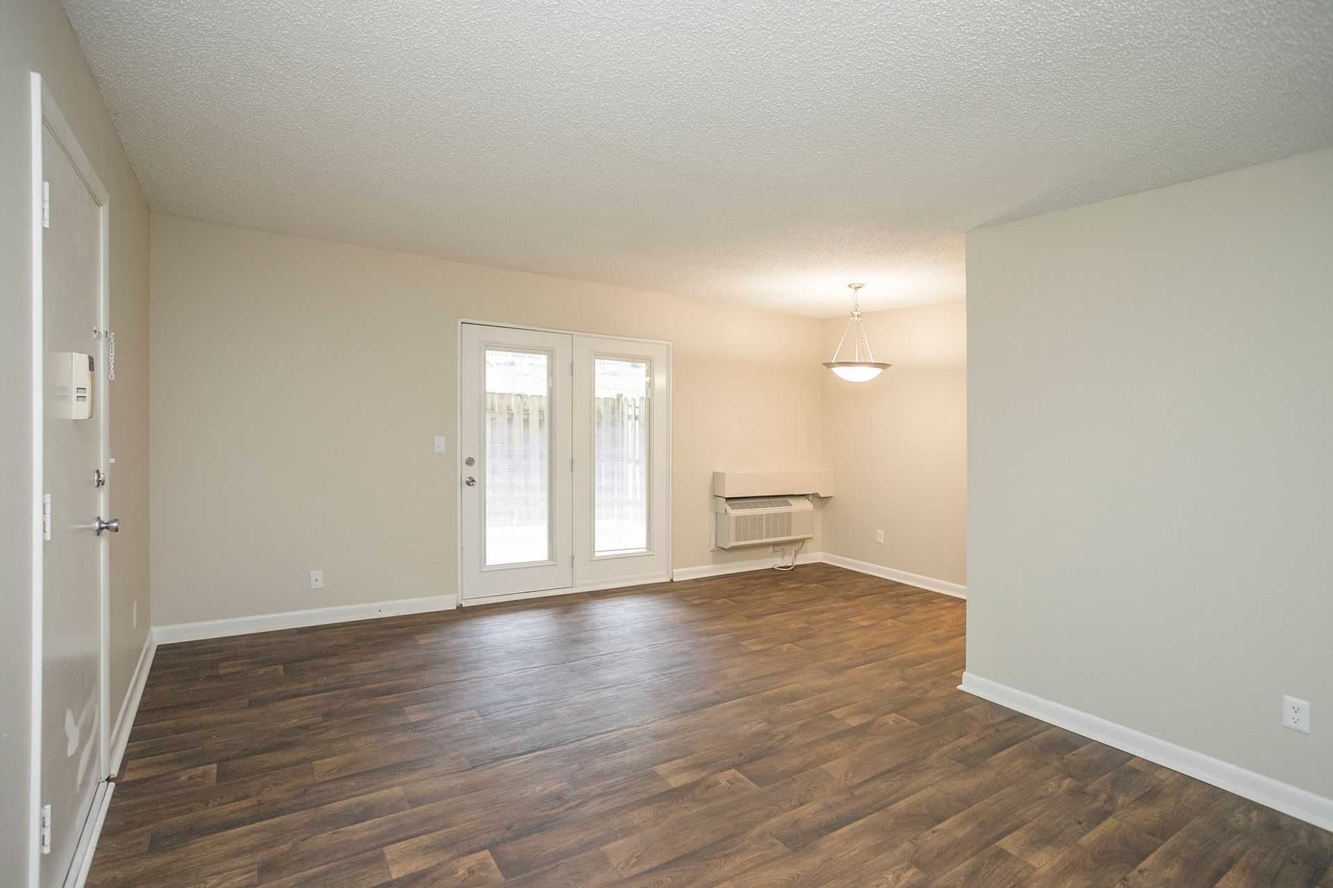 An empty room with neutral walls, wood-look flooring, a glass sliding door, and a light fixture hanging in the back.