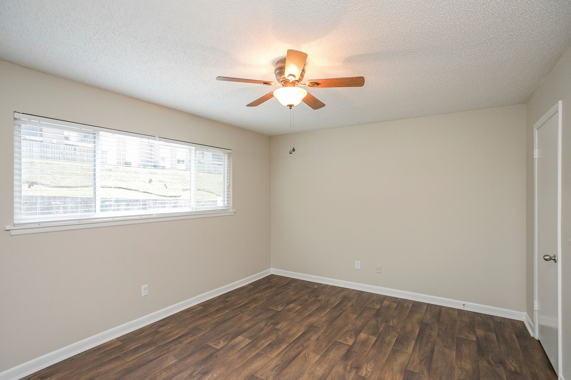 An empty, neutral-toned room featuring light wood floors, beige walls, a white-trimmed window, and a ceiling fan.