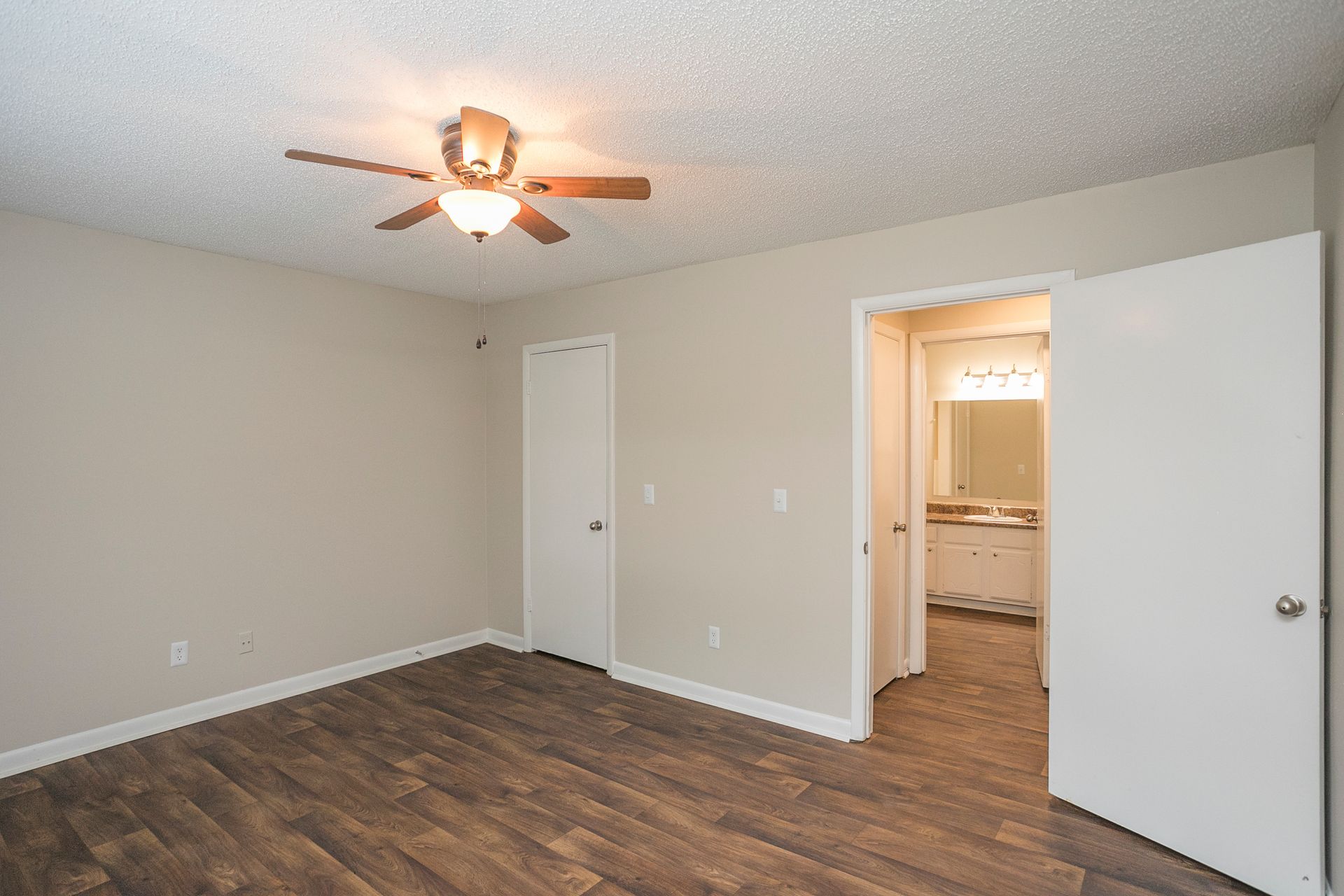 Empty bedroom with wood-look flooring, tan walls, a ceiling fan, a closed closet door, and an open door to a bathroom.