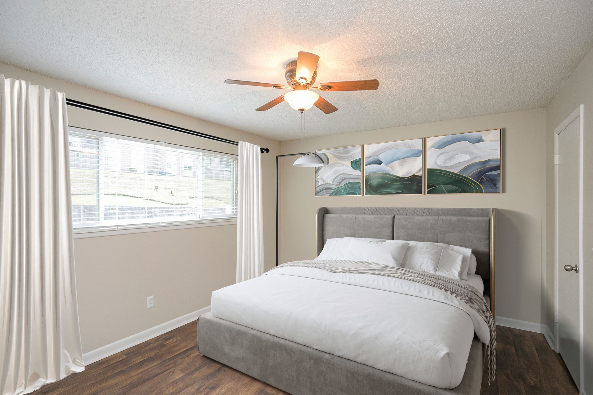 A cozy bedroom featuring a grey upholstered bed, light walls, wooden floors, a ceiling fan, and a wide window with curtains.