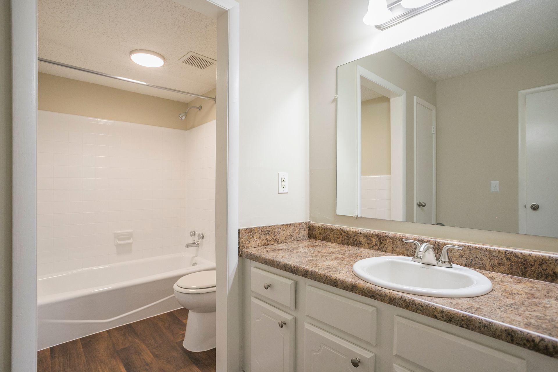 A bathroom featuring a granite-topped vanity with a white sink, a large mirror, and an adjoining room with a tub and toilet.