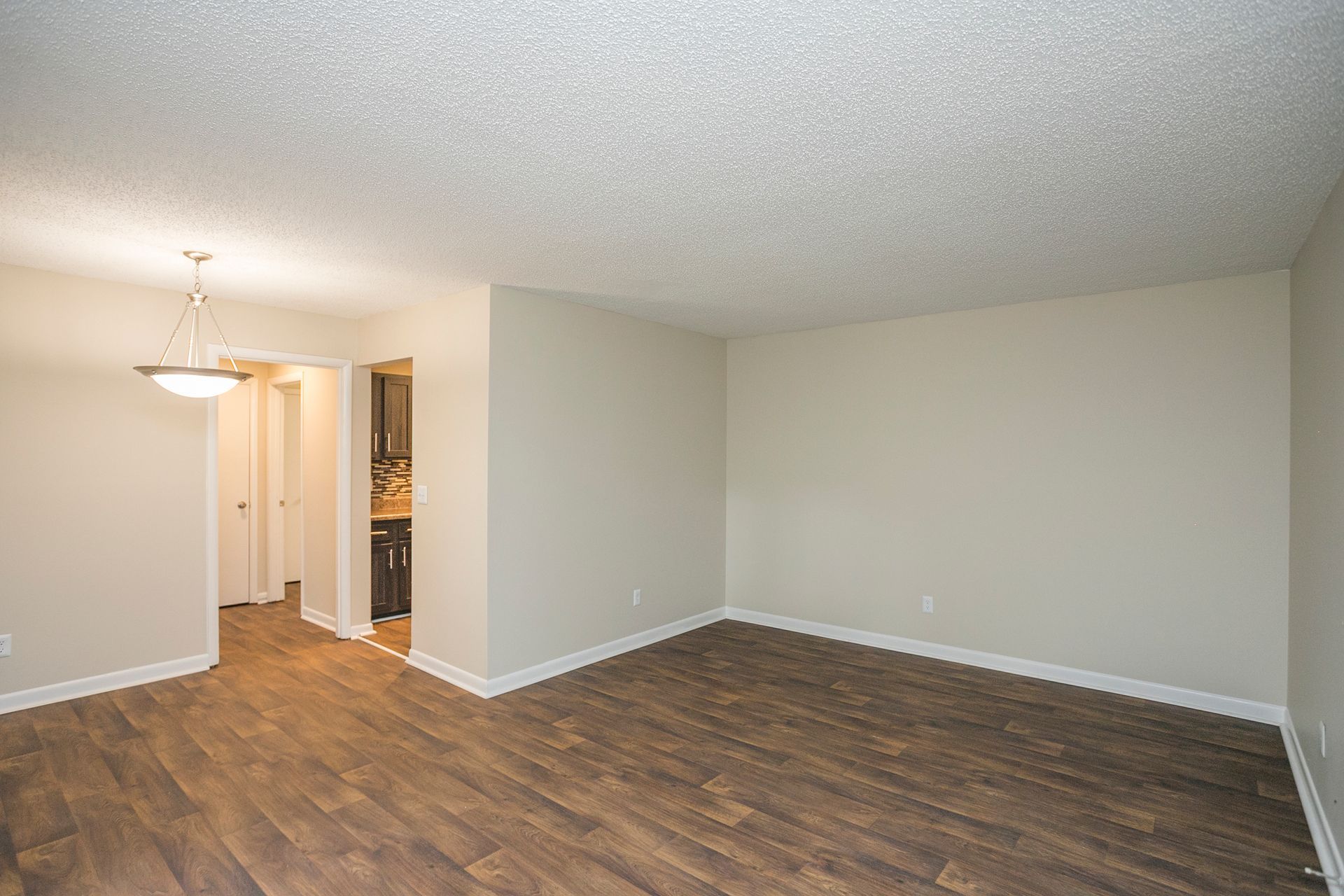 An empty, beige-walled living room with wood-look flooring and a light fixture, leading into a kitchen area.