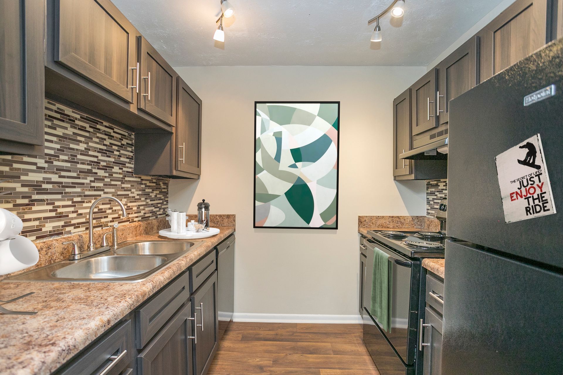 A narrow galley kitchen with dark wood cabinets, tan granite counters, tile backsplash, and a black refrigerator.