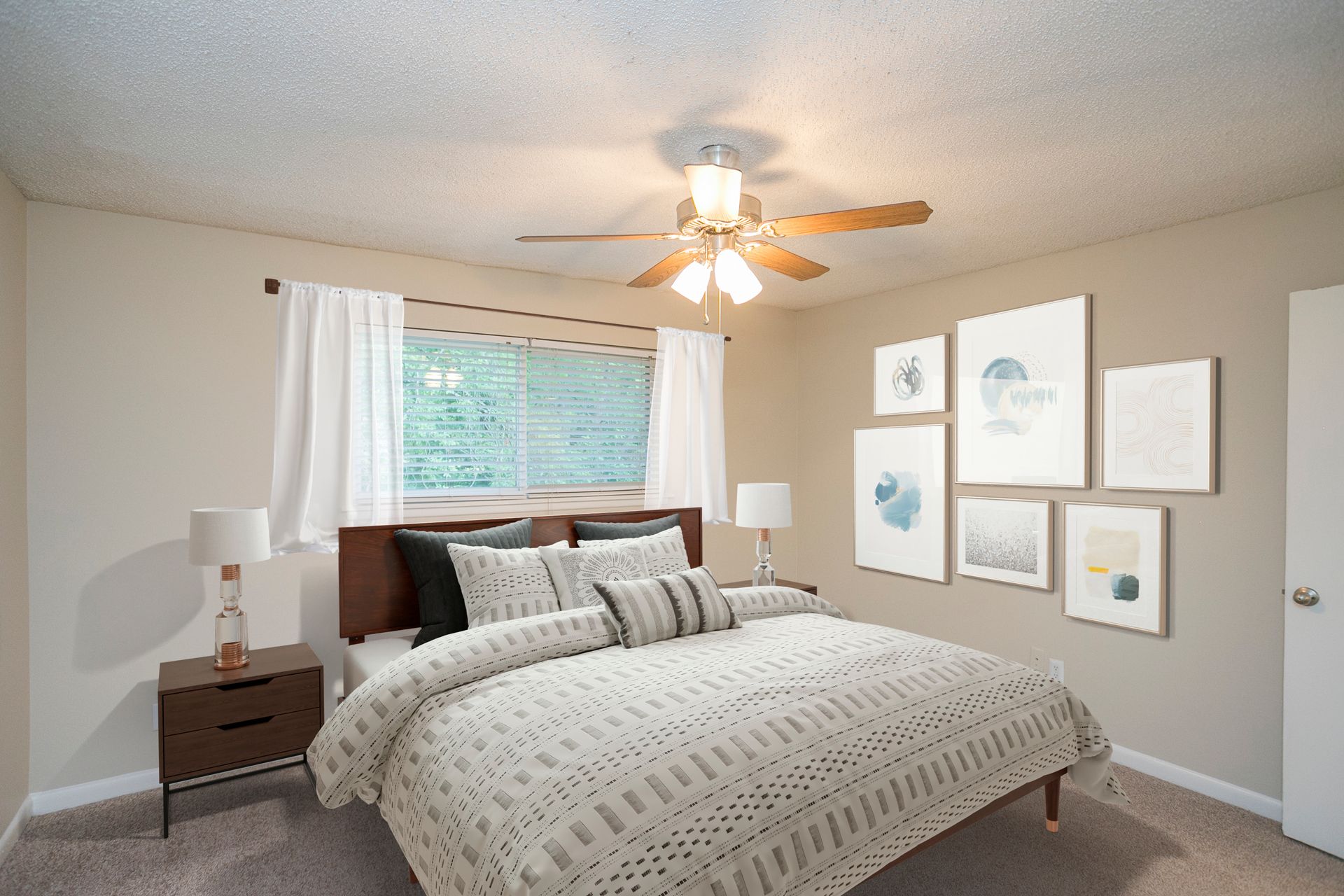 A bedroom featuring a bed with patterned bedding, a nightstand with a lamp, white curtains, and wall art in a grid.