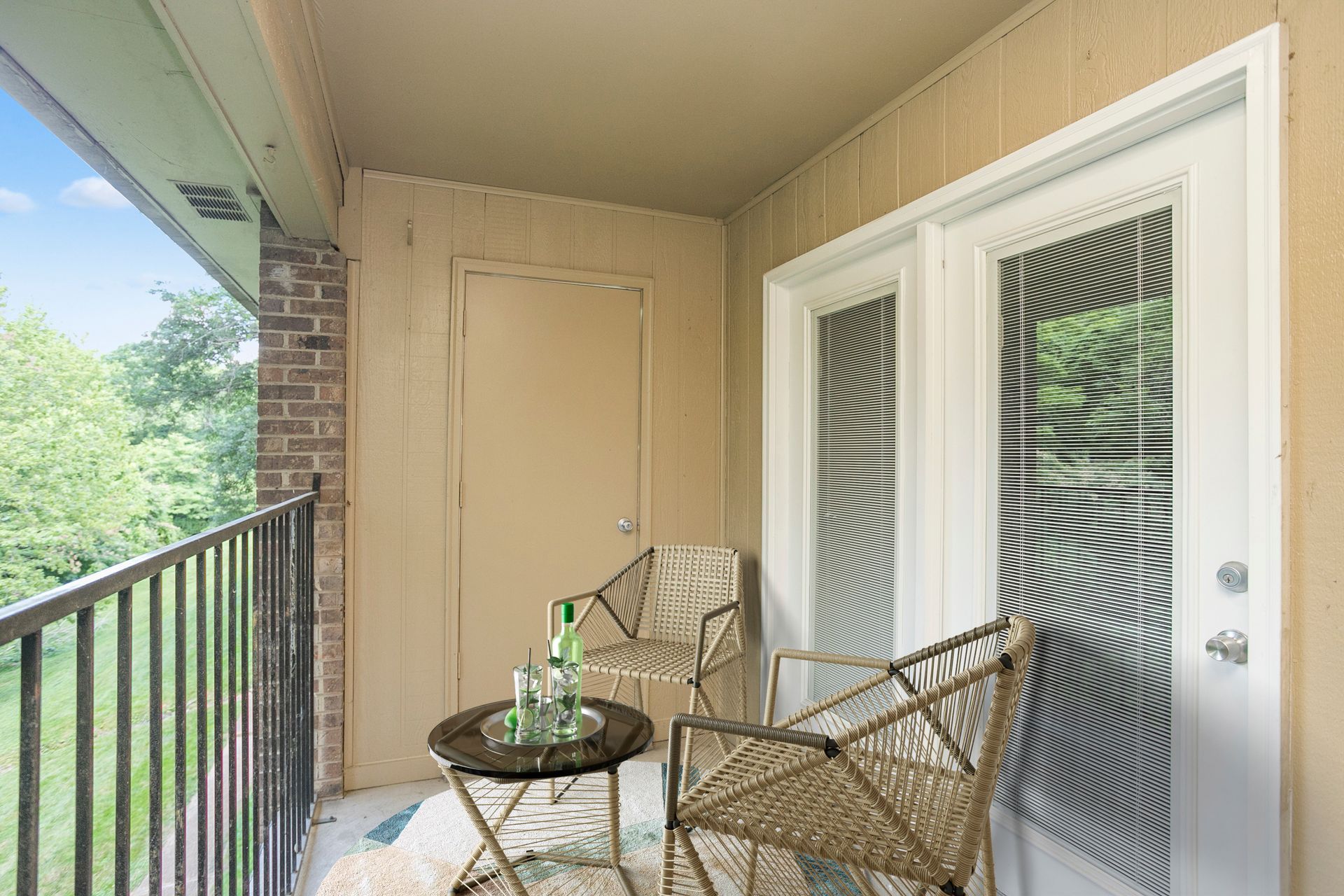 A balcony with a small round table, two woven chairs, and a glass door overlooking a leafy outdoor area.