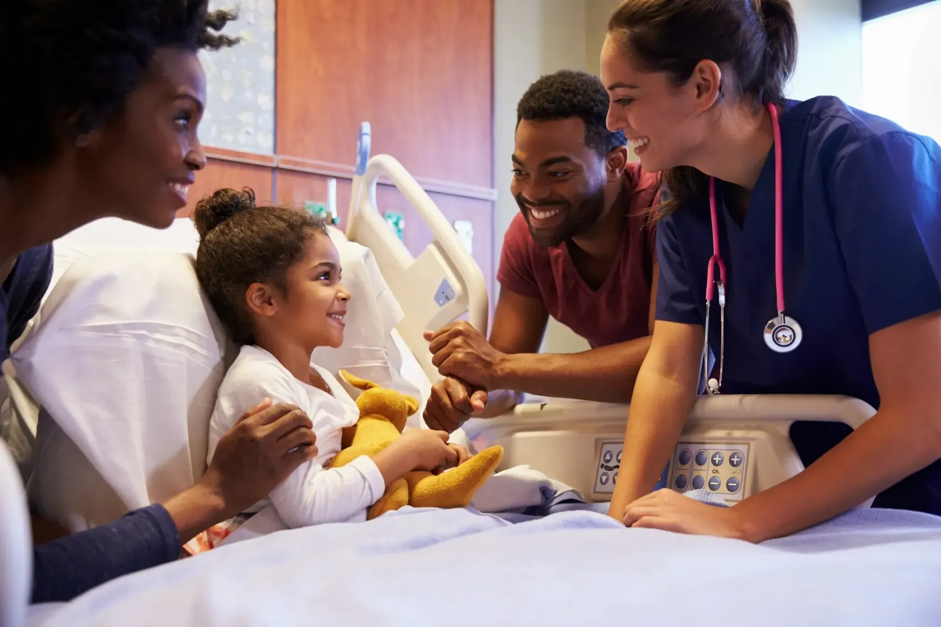 Healthcare worker smiling with family beside a hospital bed, supporting a child during care
